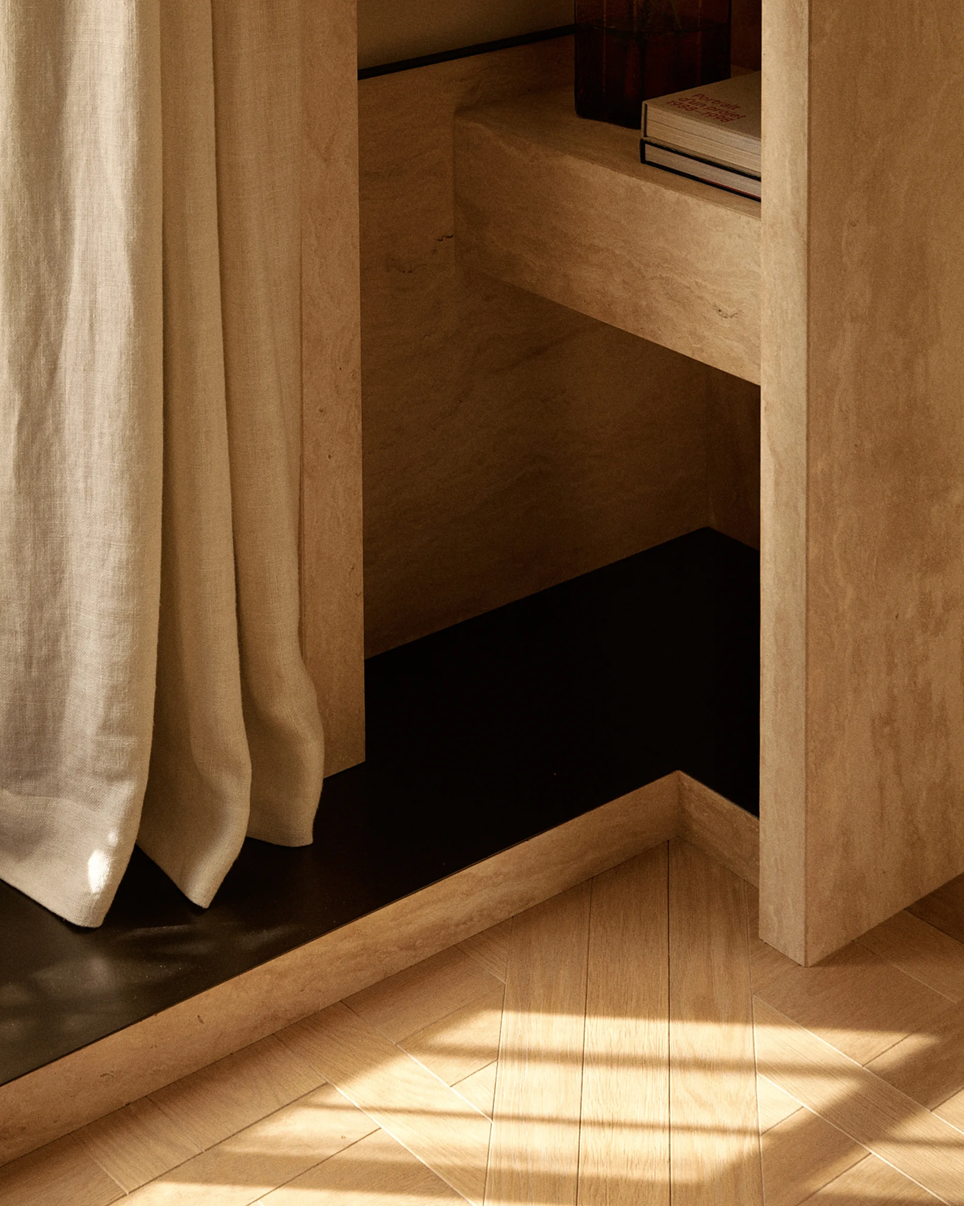A close-up of a stone-textured built-in shelf unit with books next to a linen curtain and herringbone pattern wood flooring with soft sunlight.
