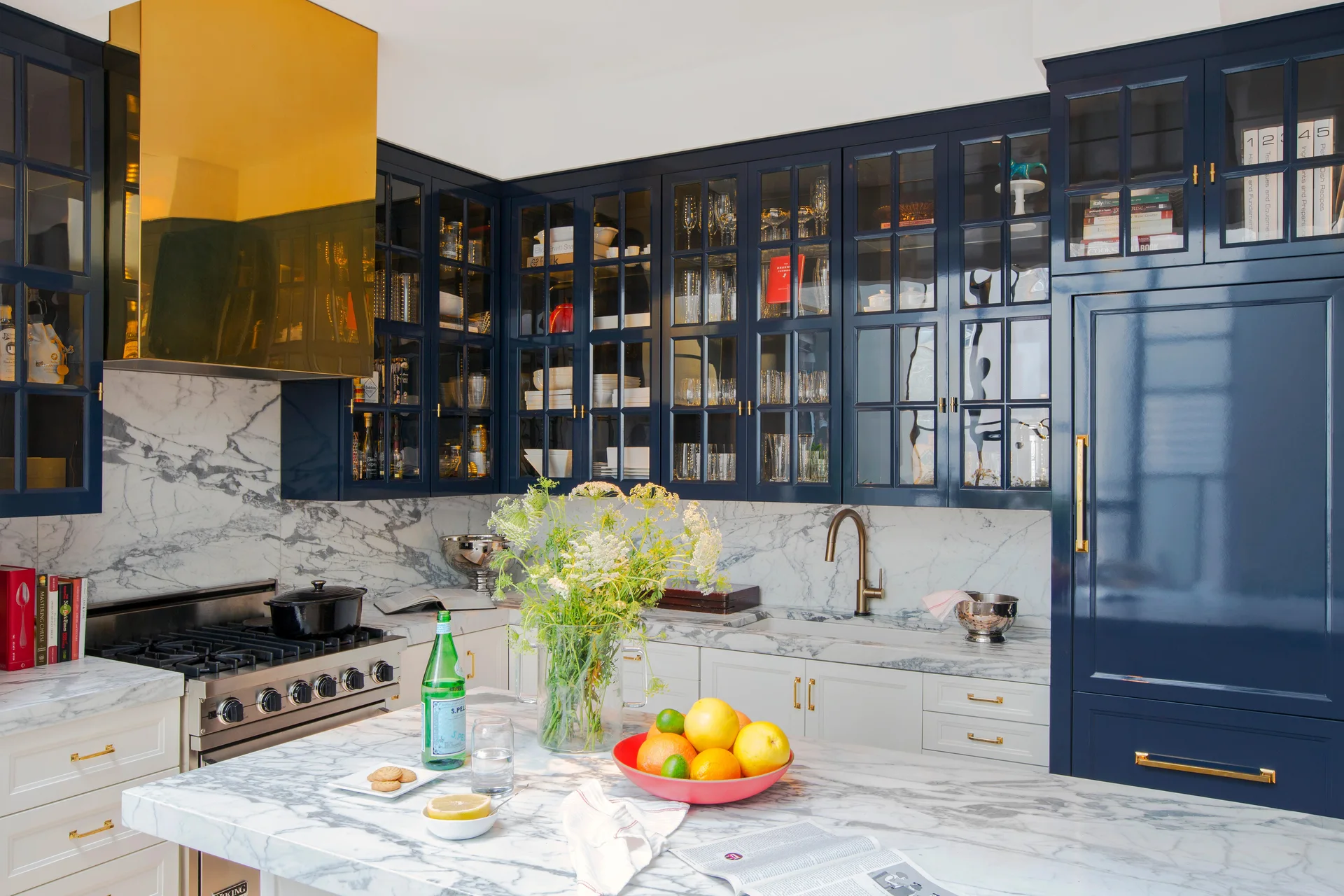 A kitchen with navy blue upper cabinets, white lower cabinets, a veined marble backsplash, a brass range hood, and a marble-topped island.