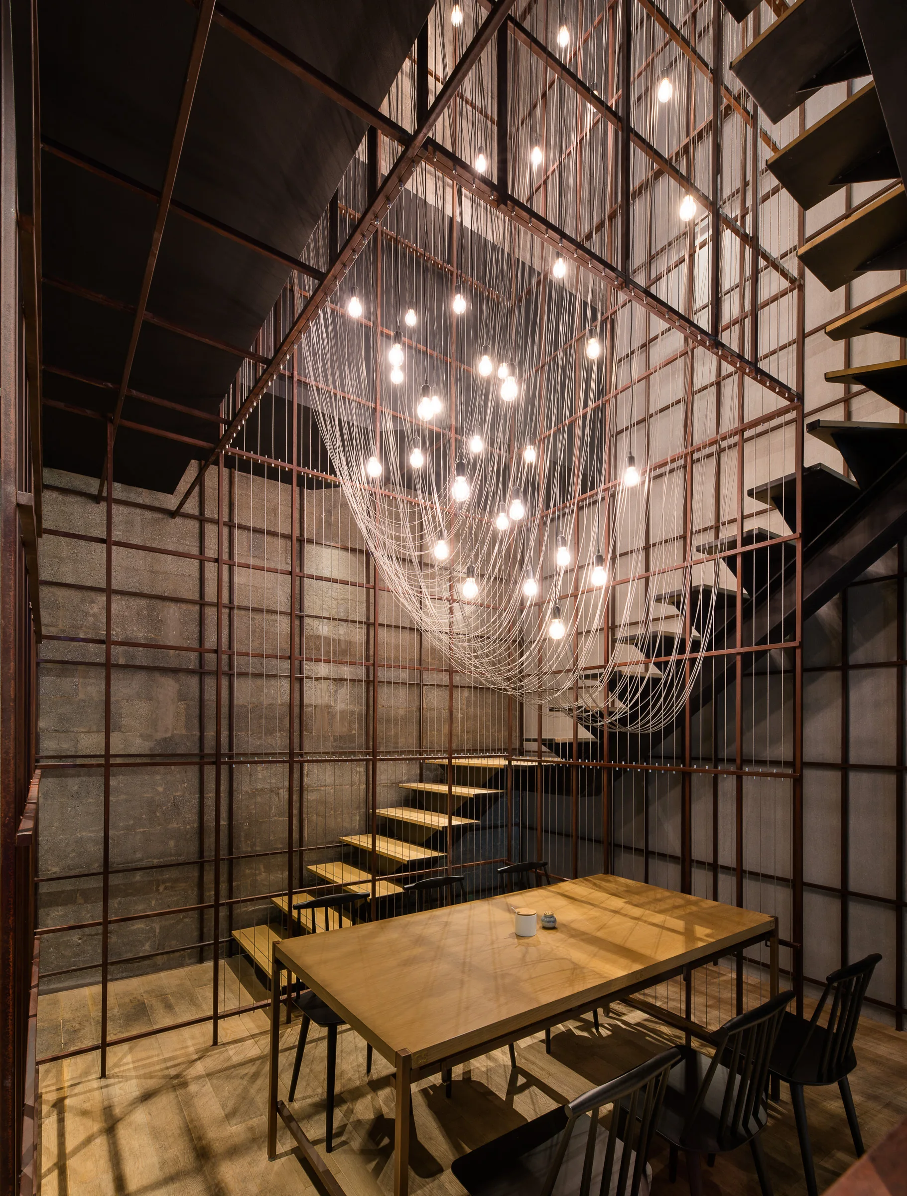 An industrial dining space with a wood table and black chairs enclosed in a tall metal grid under a sweeping cluster of hanging light bulbs.