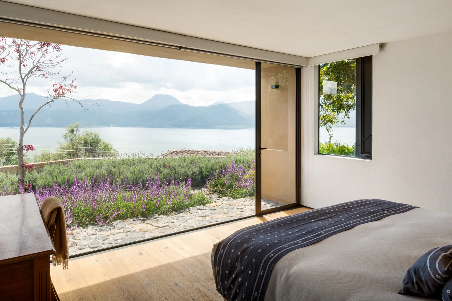 A minimalist bedroom featuring light wood flooring, off-white walls, and a large sliding glass door that opens to a garden and lake view.