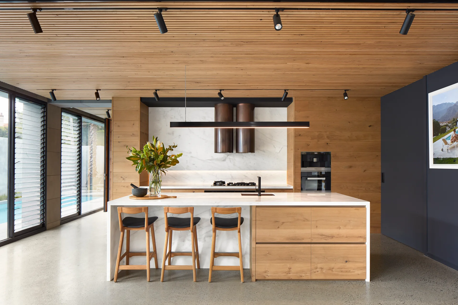 A modern kitchen features a central marble-topped island with wooden stools, a wood-slatted ceiling, and integrated wooden cabinetry.