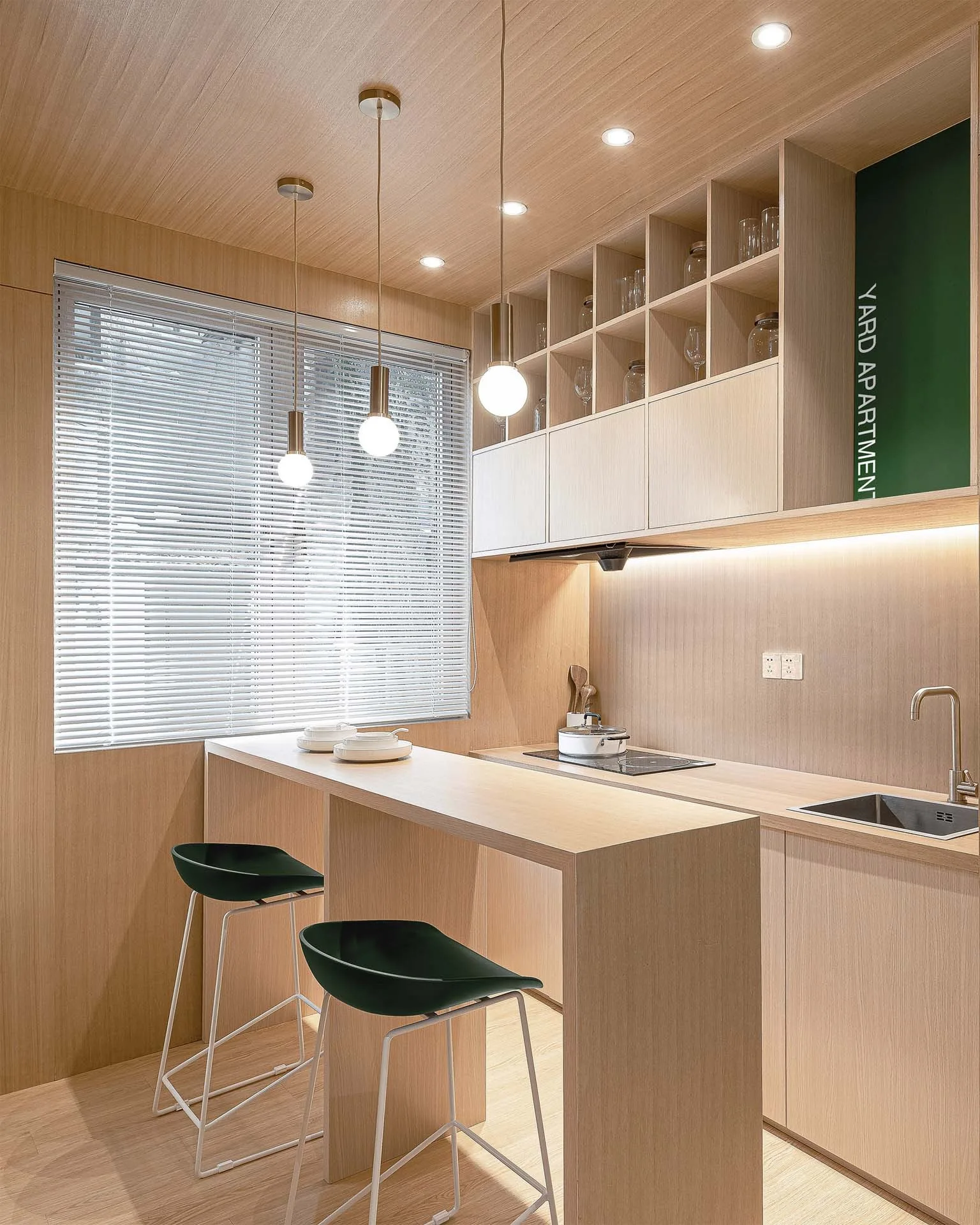 A compact modern kitchen wrapped in light wood paneling, featuring a matching breakfast bar peninsula with dark green stools and globe pendant lights.