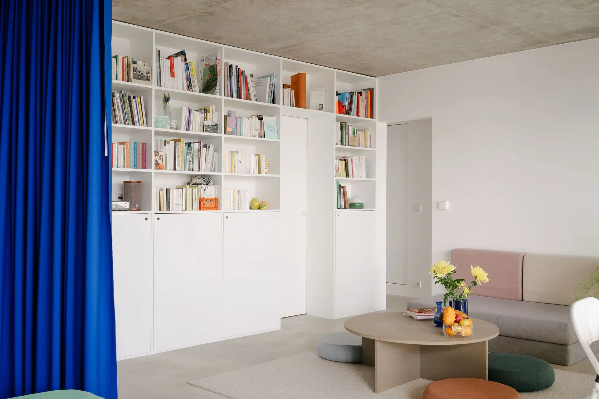 A minimalist living room features a white built-in bookshelf wall, a raw concrete ceiling, a low round coffee table, and a vibrant blue curtain.
