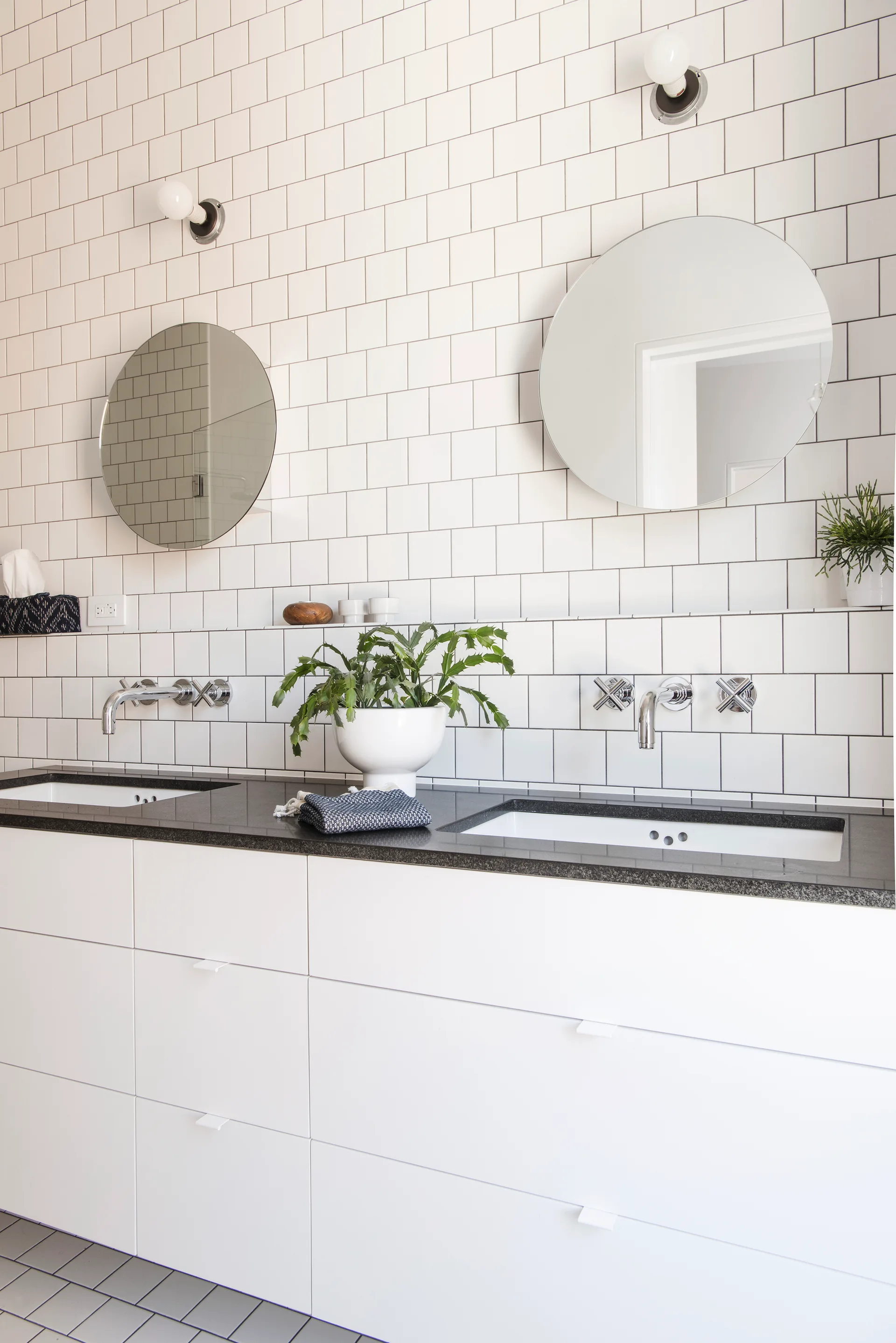 A modern double bathroom vanity featuring flat white cabinets, a dark countertop, round mirrors, and wall-mounted faucets against a white tiled wall.