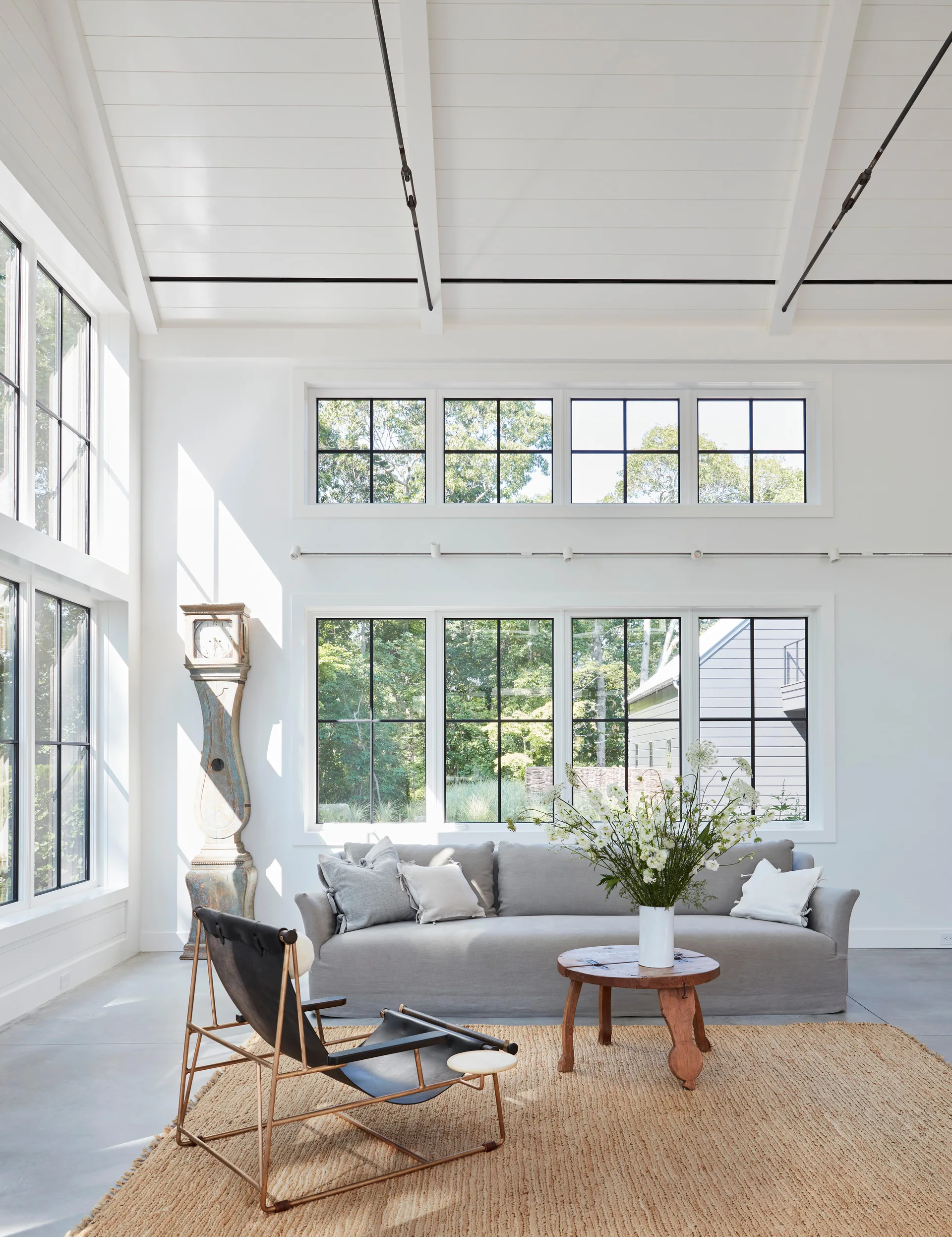 A sunlit living room features a grey sofa, a black sling chair on a jute rug, and a tall grandfather clock beneath high, vaulted ceilings.