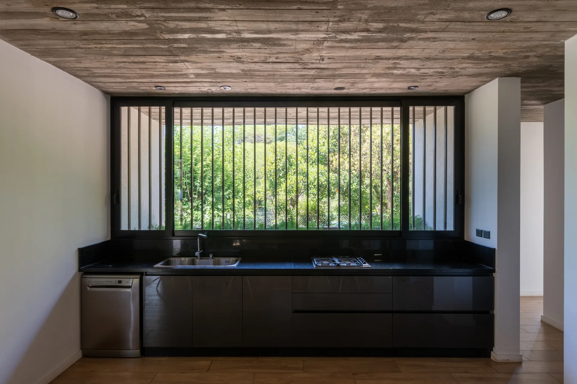A minimalist dark kitchen featuring a raw board-formed concrete ceiling, sleek dark cabinetry, and a large window with vertical metal security bars.