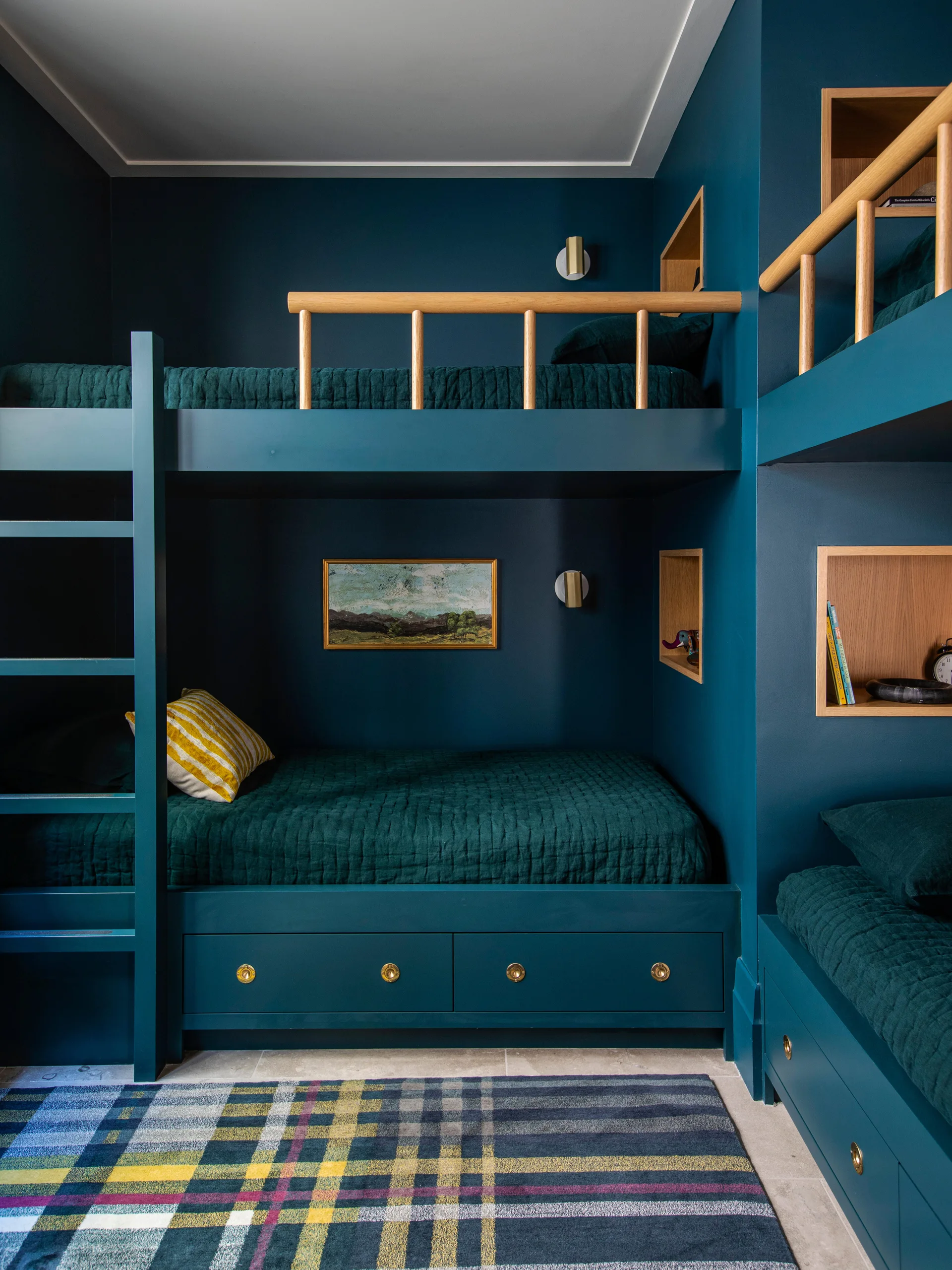Custom dark blue built-in bunk beds feature warm oak railings and recessed wooden niches next to a plaid area rug.