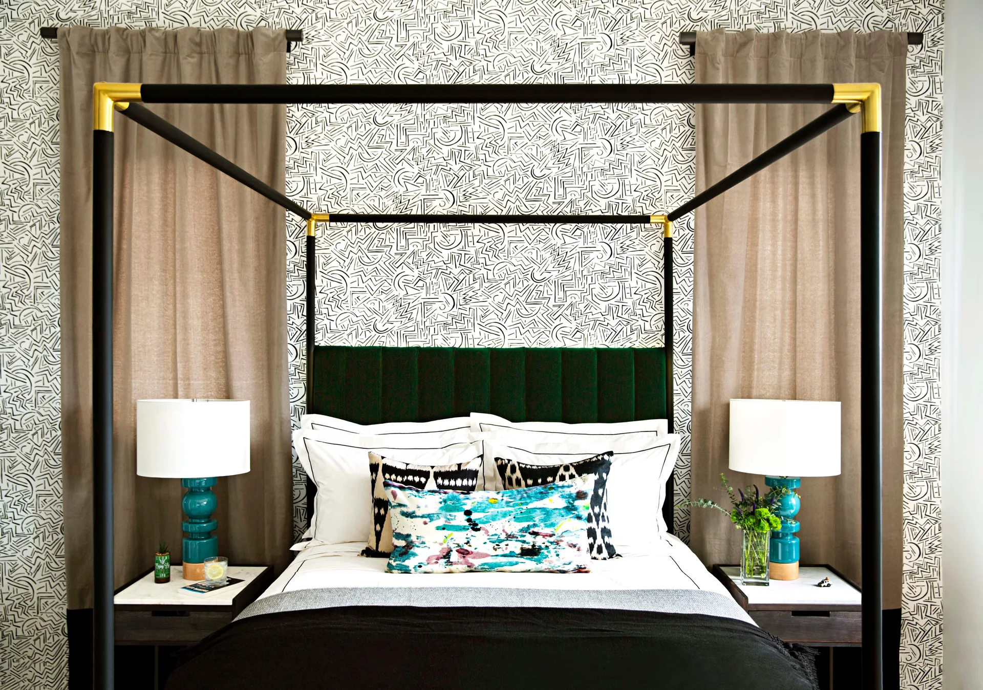 A black canopy bed with a green headboard is flanked by bright teal ceramic lamps against graphic geometric wallpaper.