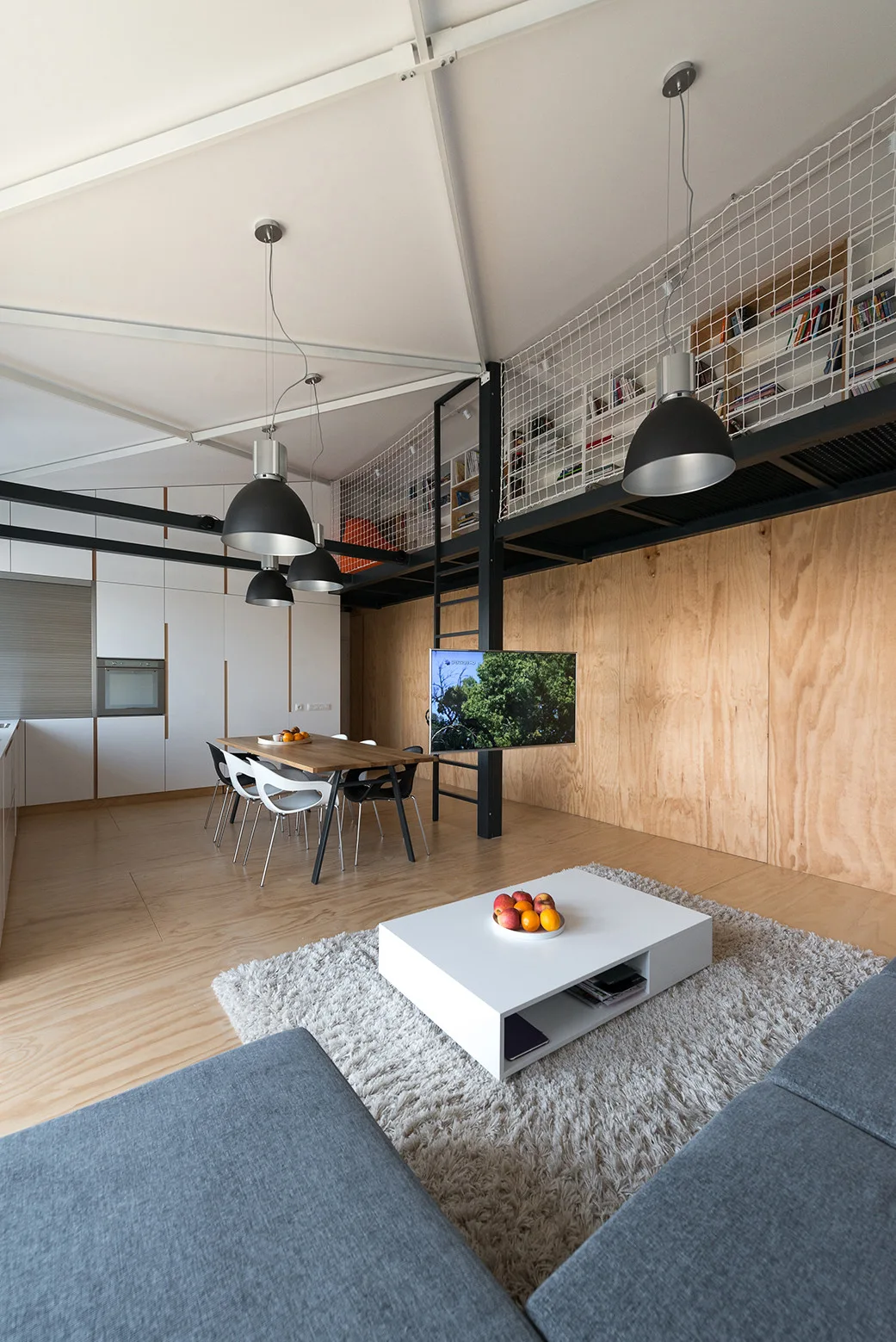 A modern loft interior with a grey sofa, a low white coffee table, a wooden dining set, and a mezzanine level with a safety net railing.
