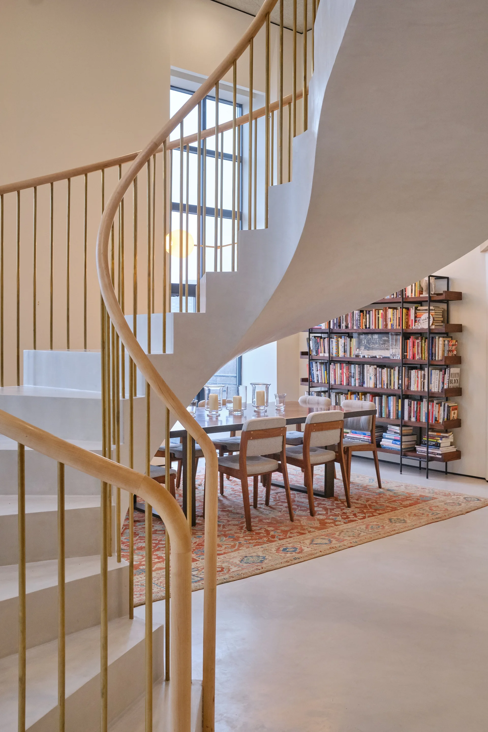 A white helical staircase with gold railings winds above a dining area with a wooden table, grey chairs, a patterned rug, and a tall bookshelf.