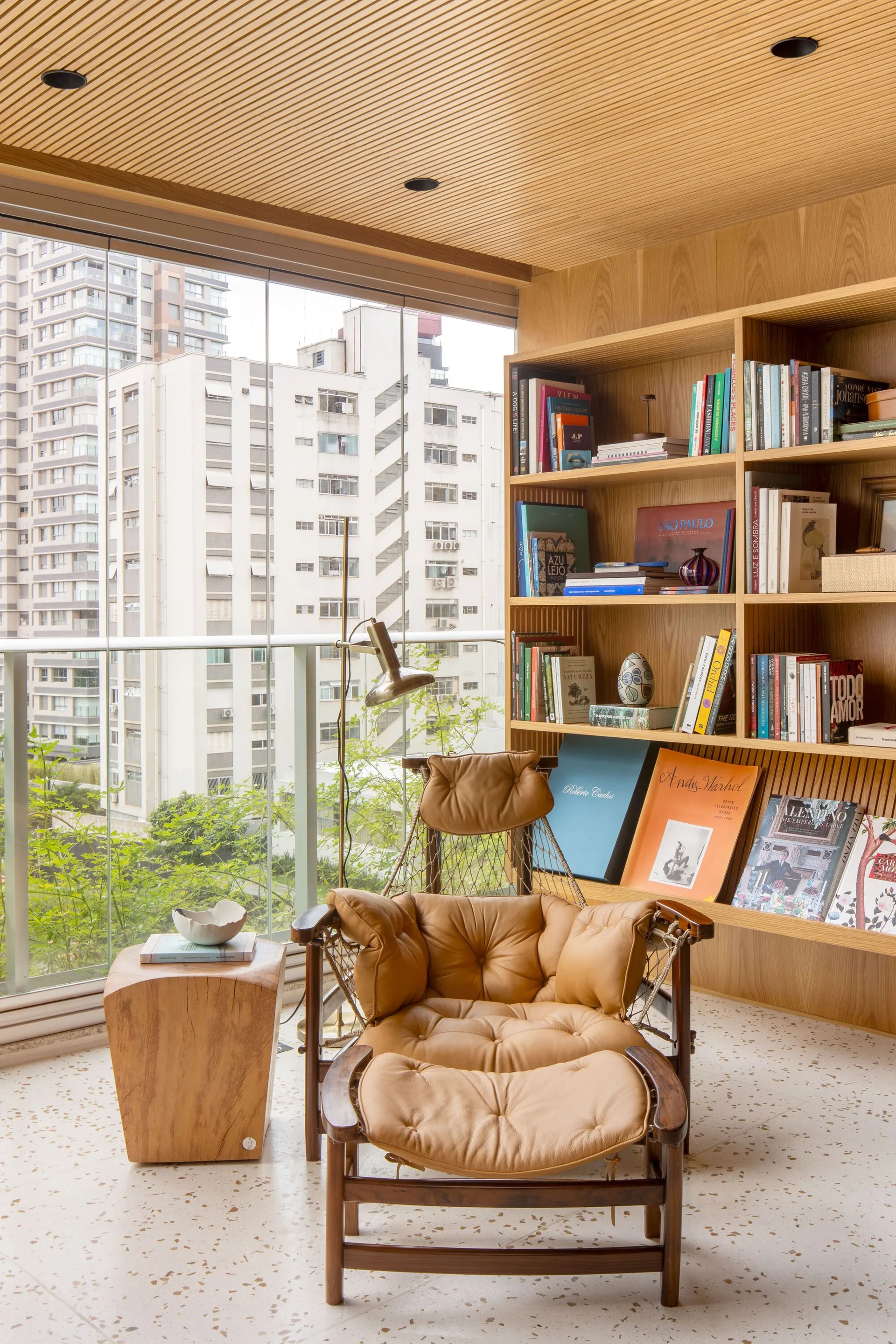 A reading corner features a tufted tan leather armchair next to a wooden bookshelf, with a floor-to-ceiling glass wall showing city views.