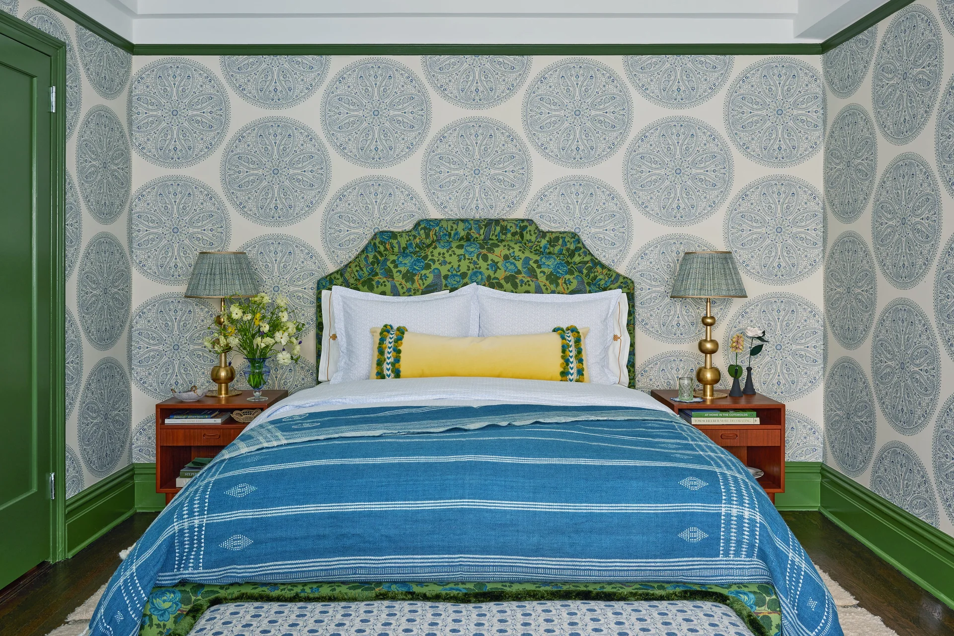 Symmetrical bedroom featuring a bed with a floral green headboard, blue medallion wallpaper, brass table lamps, and deep green trim.