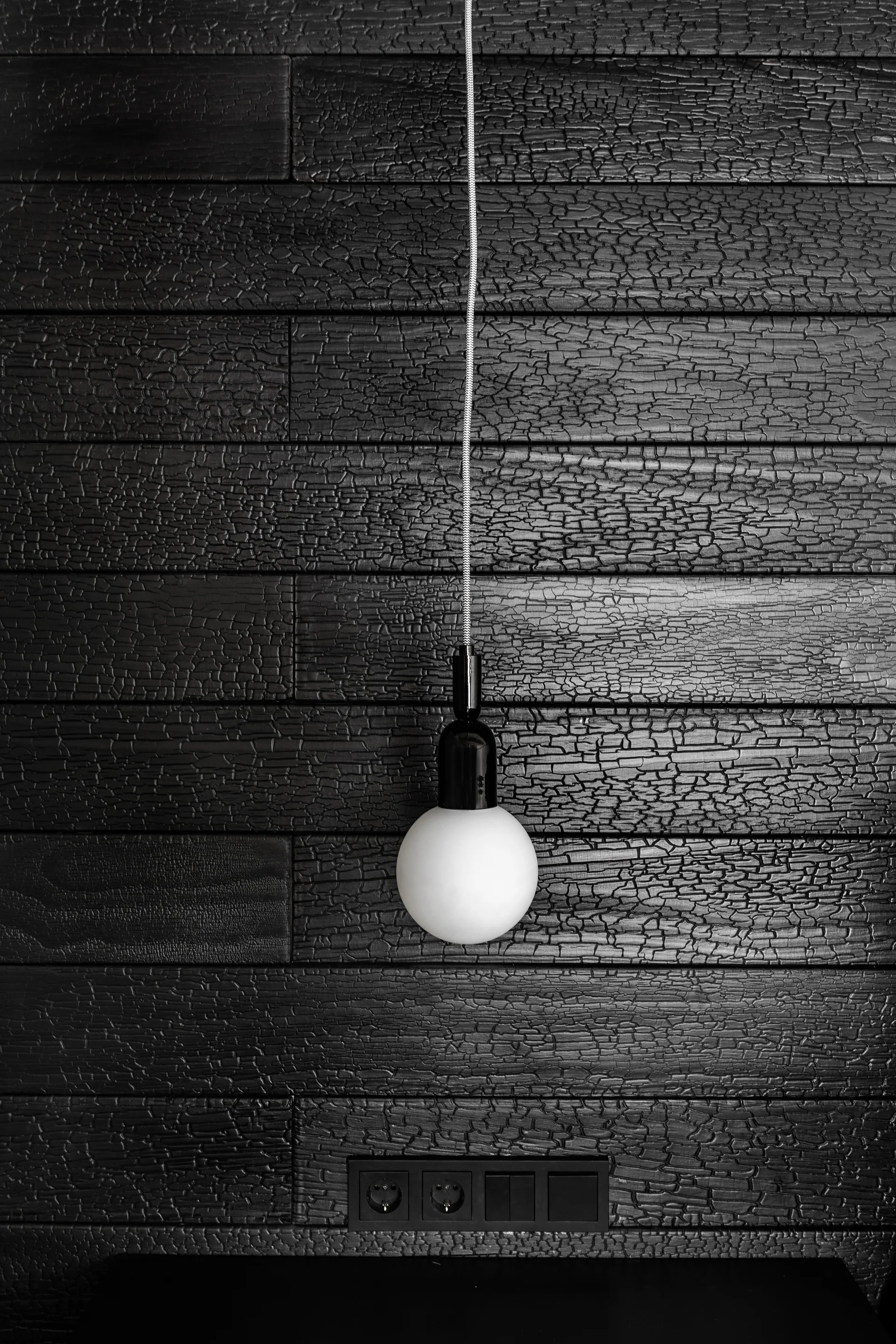 A white glass globe pendant light hangs against a black shou sugi ban charred wood wall with a matching black electrical outlet.