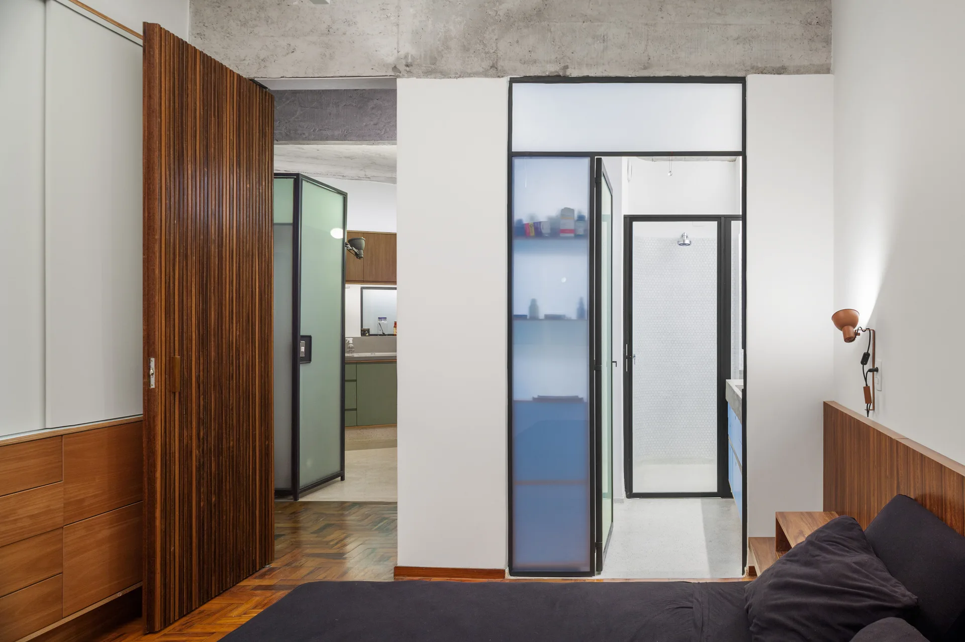 A modern bedroom featuring a dark grey bed, parquet flooring, a tall slatted wood sliding door, and a view into a bathroom with frosted glass panels.