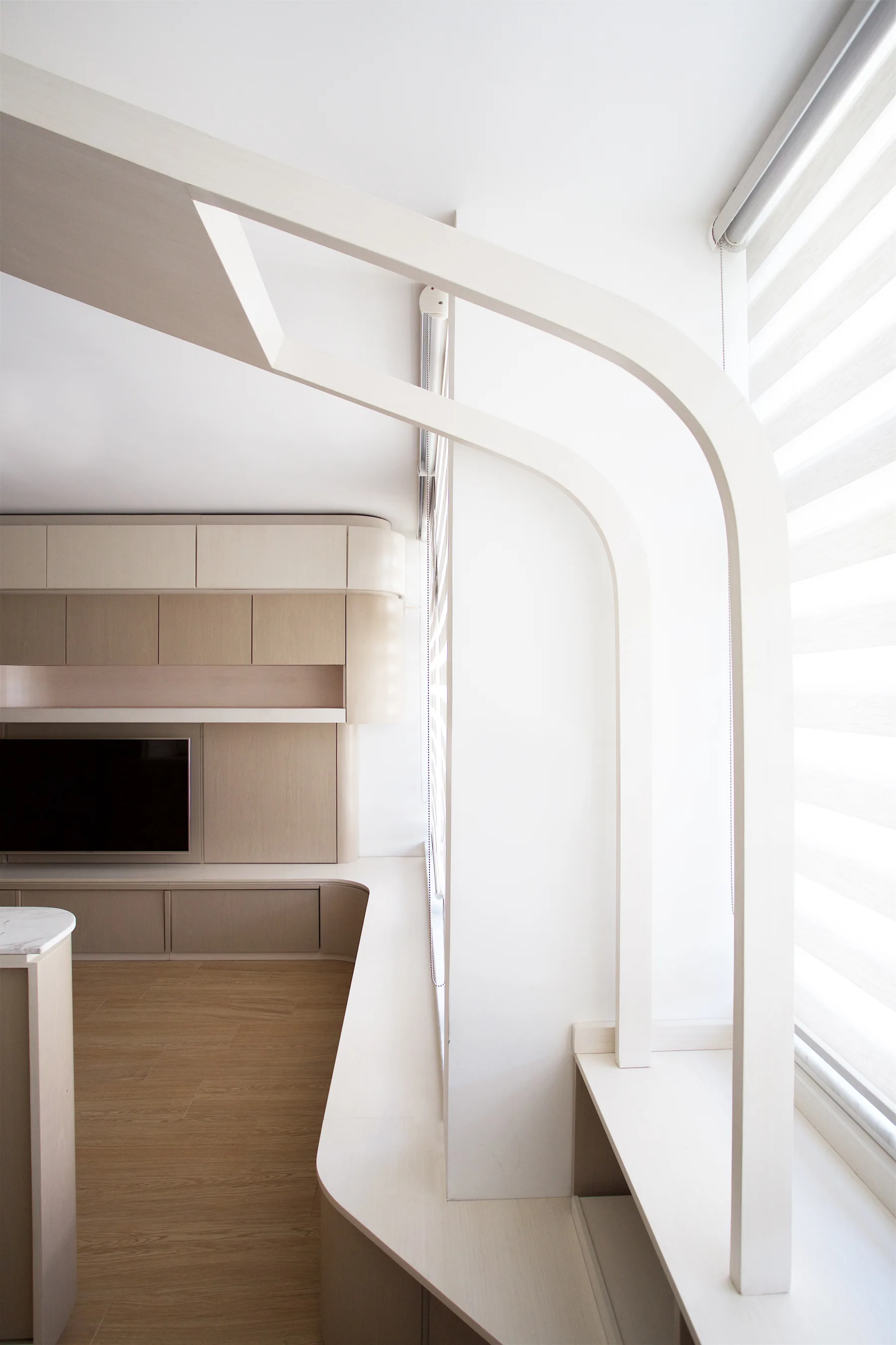 A minimalist living area features light wood built-in cabinetry with an integrated television alongside a custom curved wooden framing structure by a window.