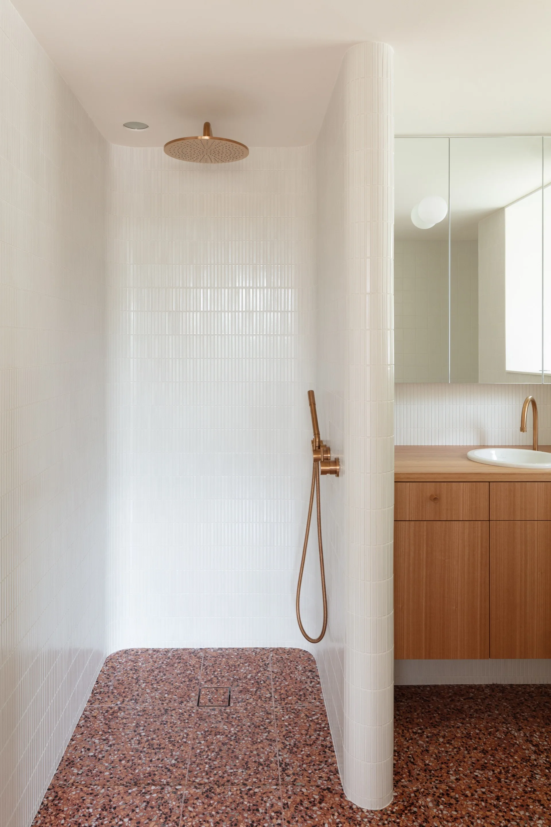 A modern bathroom features a tiled shower stall with brass fixtures next to a wood vanity, both set on a terracotta terrazzo floor.