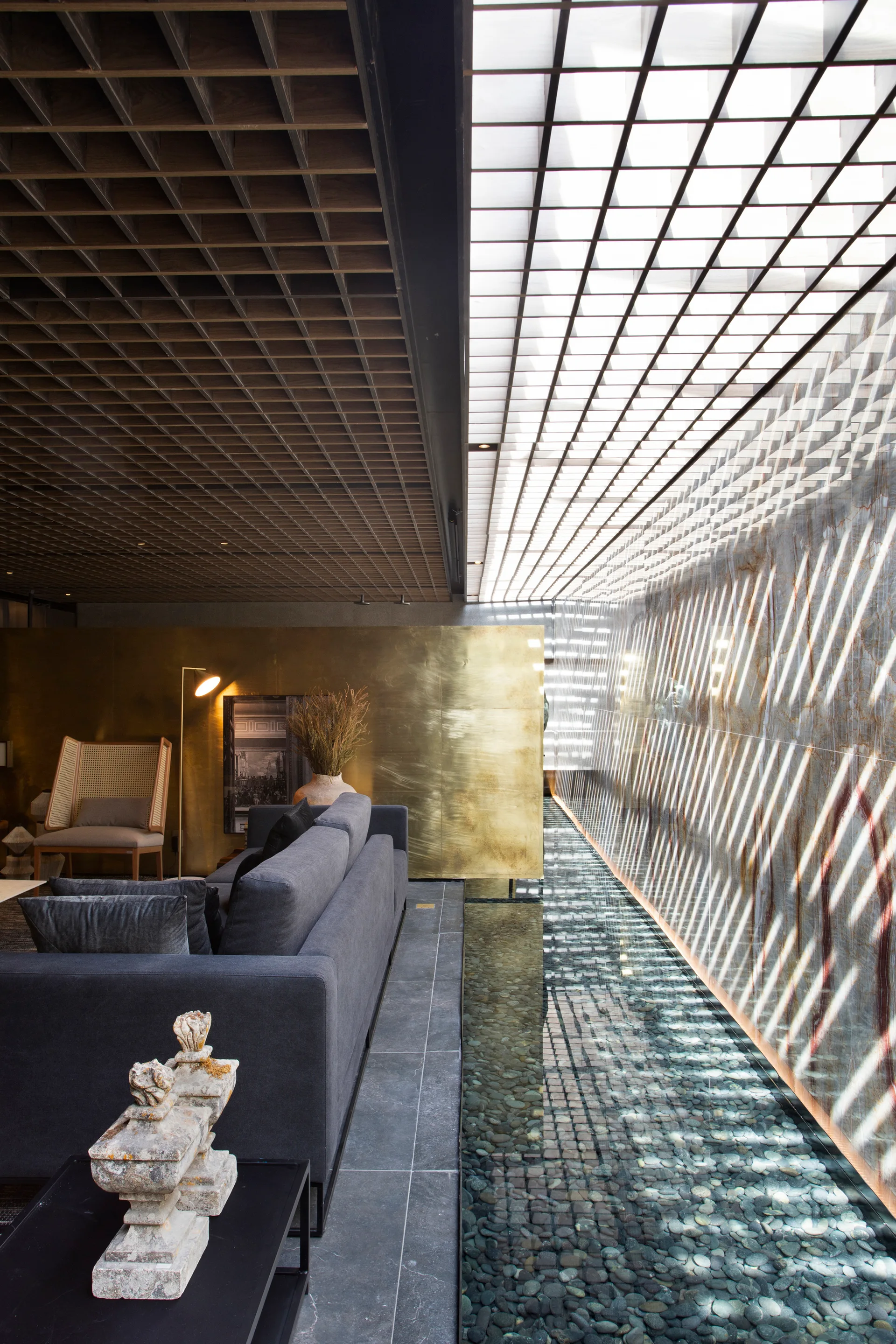 A modern living room features a grey sofa next to a long pebble-filled indoor water feature beneath a wood grid ceiling and skylight.
