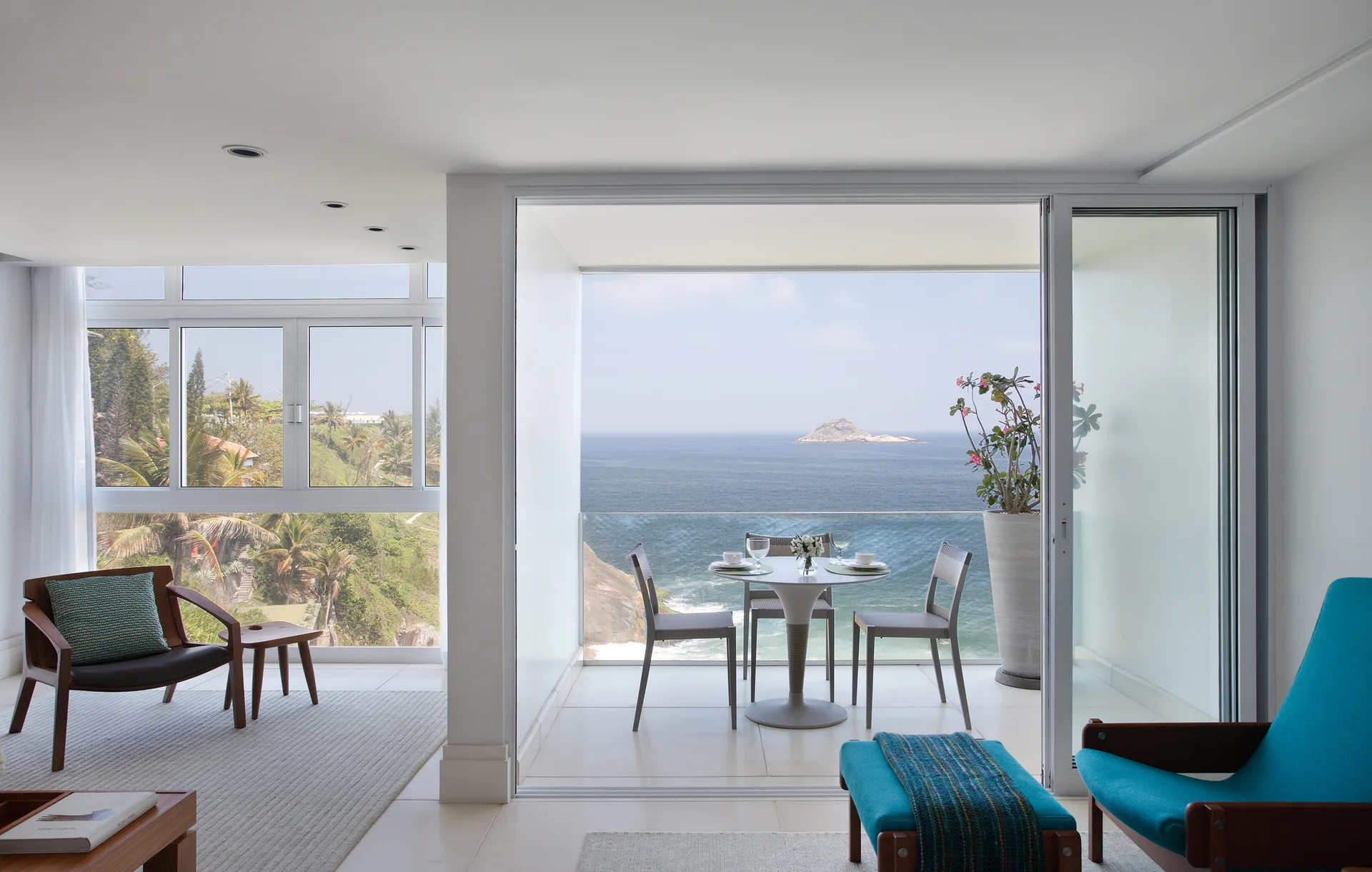 A modern living room with blue chairs opens to a tiled balcony with a dining set overlooking the blue ocean.