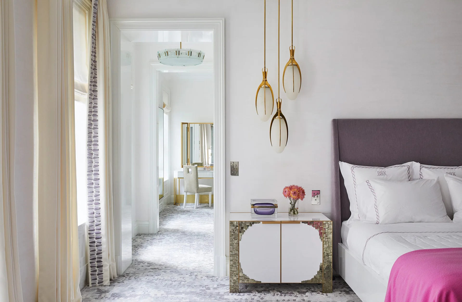 A bedroom featuring a purple upholstered headboard, a mosaic-tiled nightstand with pendant lights, and an open doorway leading to a bright dressing room.