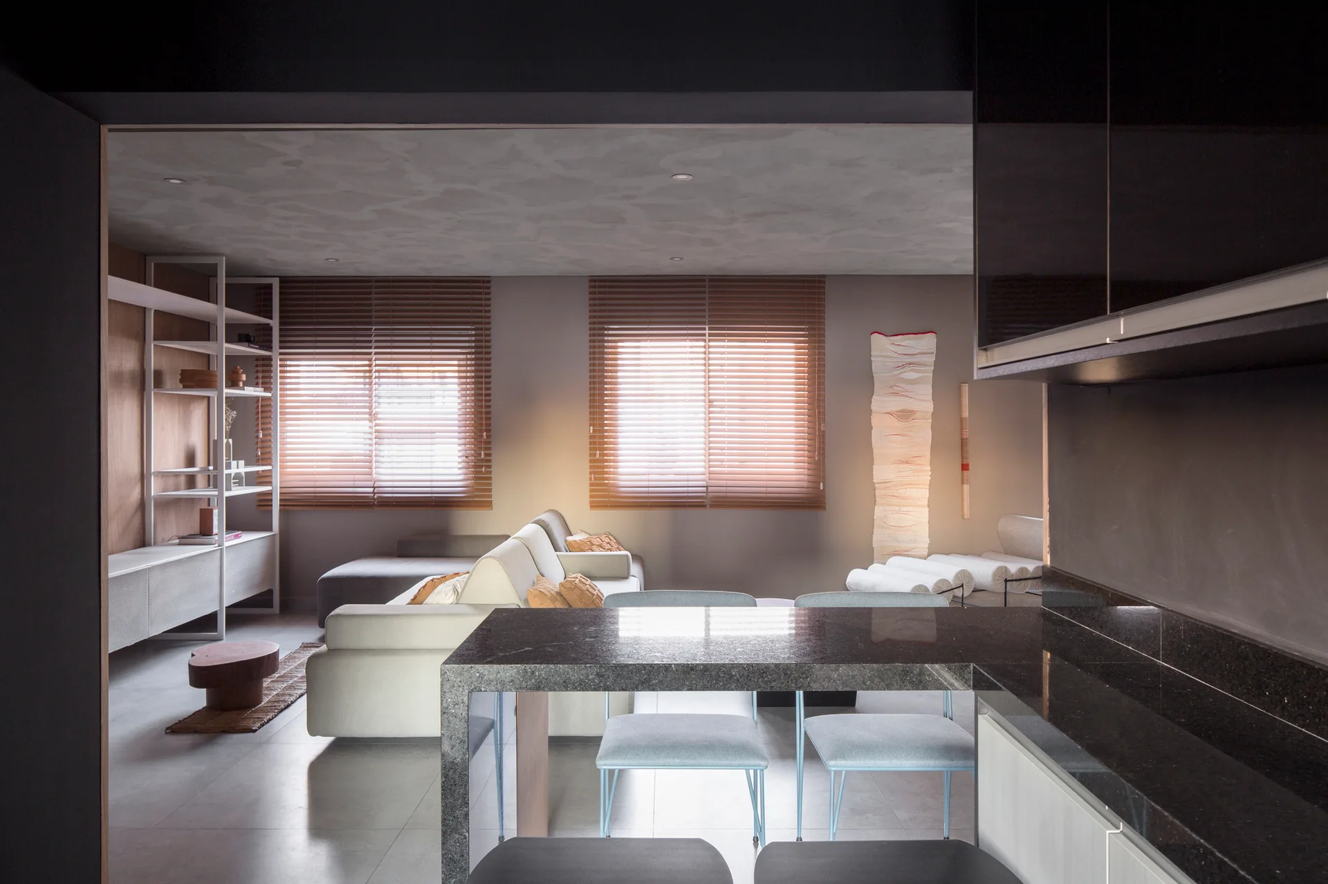 A contemporary open-plan living room and kitchen featuring a dark stone-topped island, light grey sofas, and warm wood venetian blinds on two windows.
