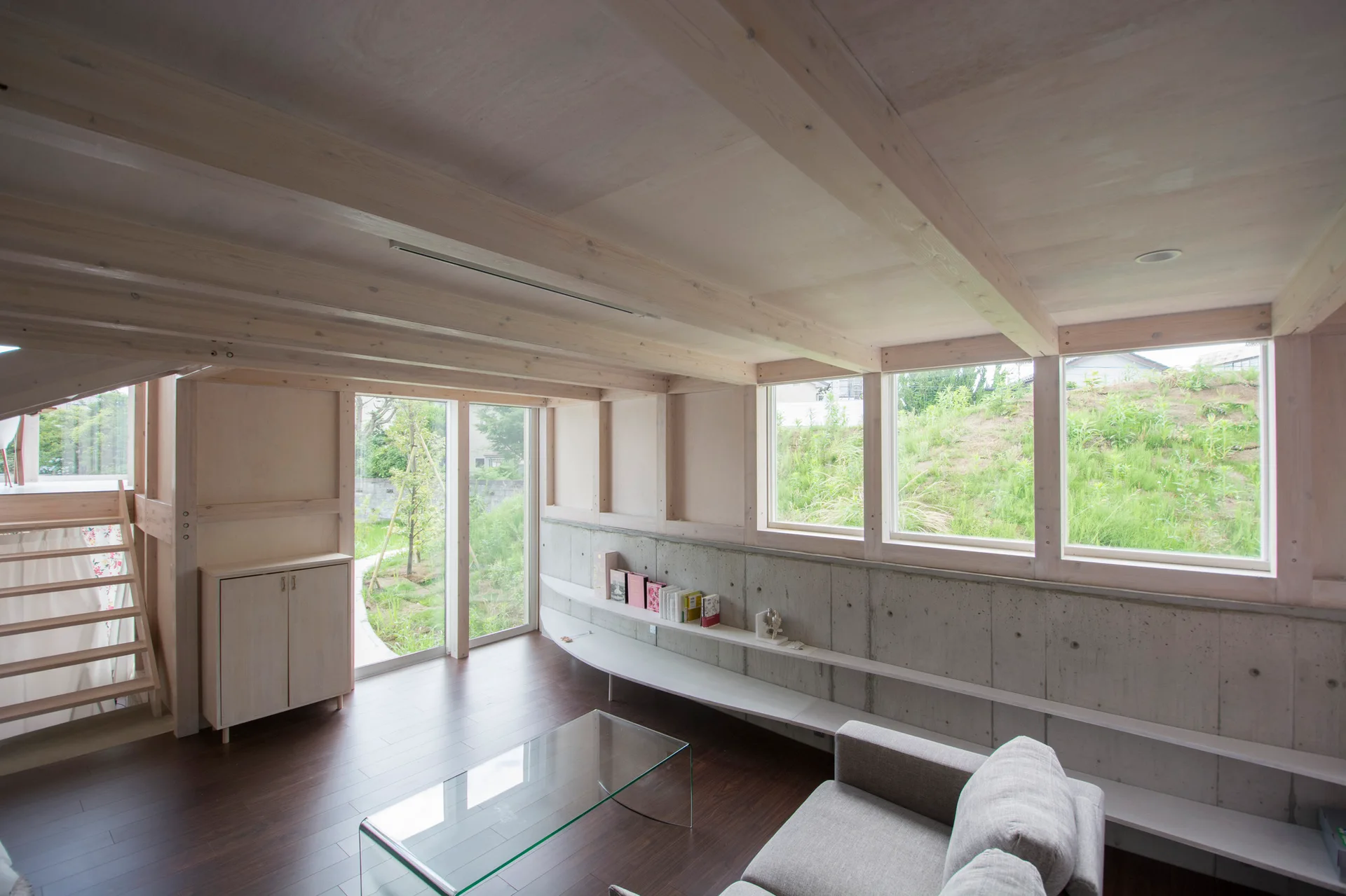 A minimalist living room features a grey sofa, a glass coffee table, and an exposed concrete wall beneath light wood ceiling beams.