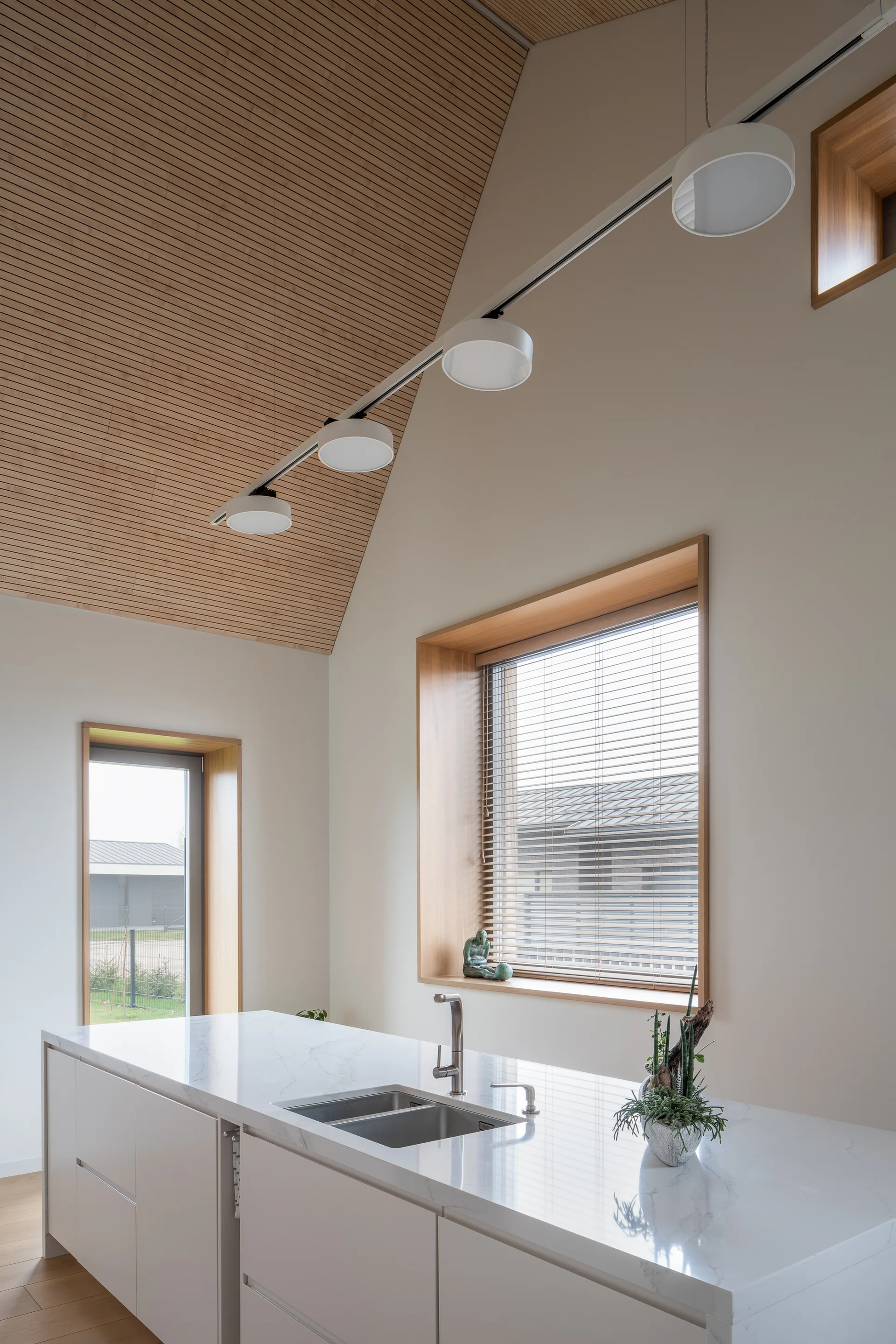 A minimalist white kitchen island with a sink sits beneath a high vaulted ceiling made of light wood slats and deep-set wooden windows.