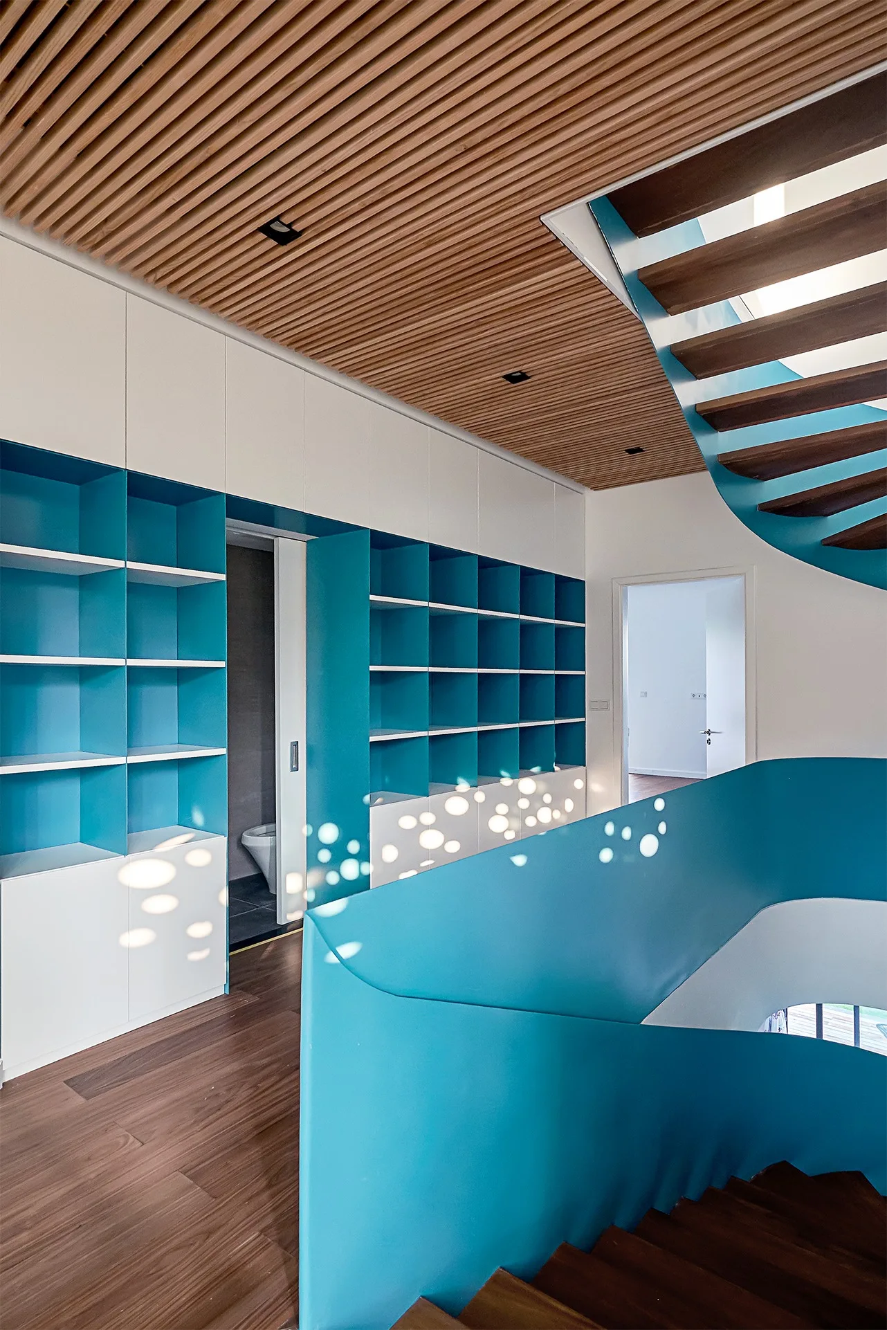 A modern hallway features a curved staircase with a bold blue railing, integrated blue shelving, and a warm wood slat ceiling.
