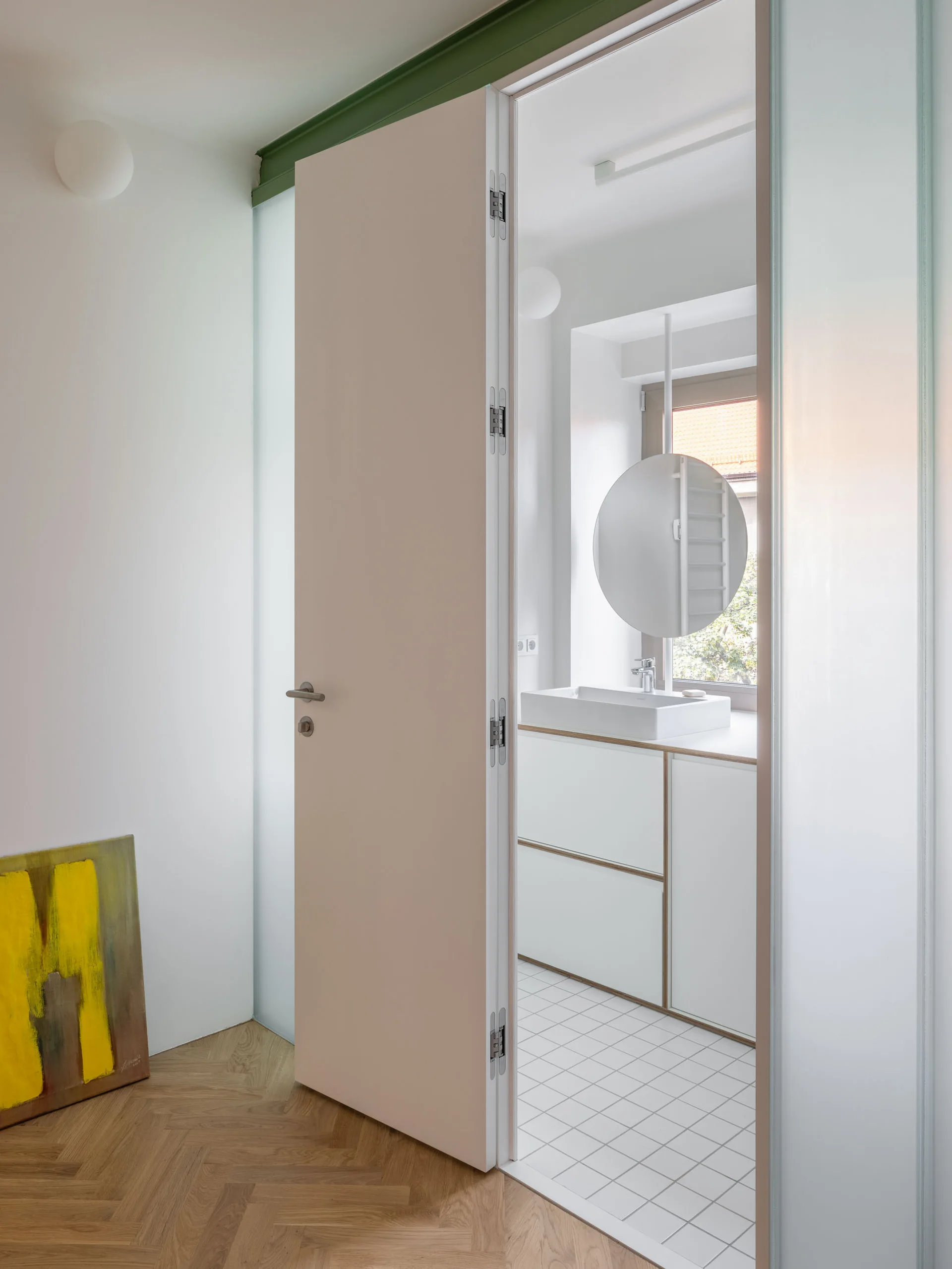 An open door reveals a minimalist bathroom featuring a white vanity, a round mirror suspended near a window, and white grid tile flooring.