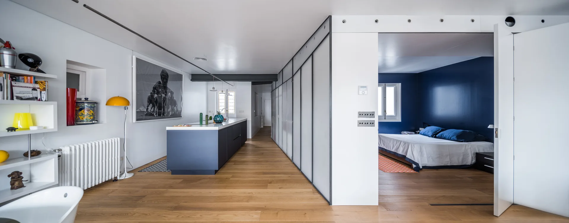 An open-concept kitchen with a navy blue island and light wood floors features a translucent sliding partition leading to a bedroom with deep blue walls.