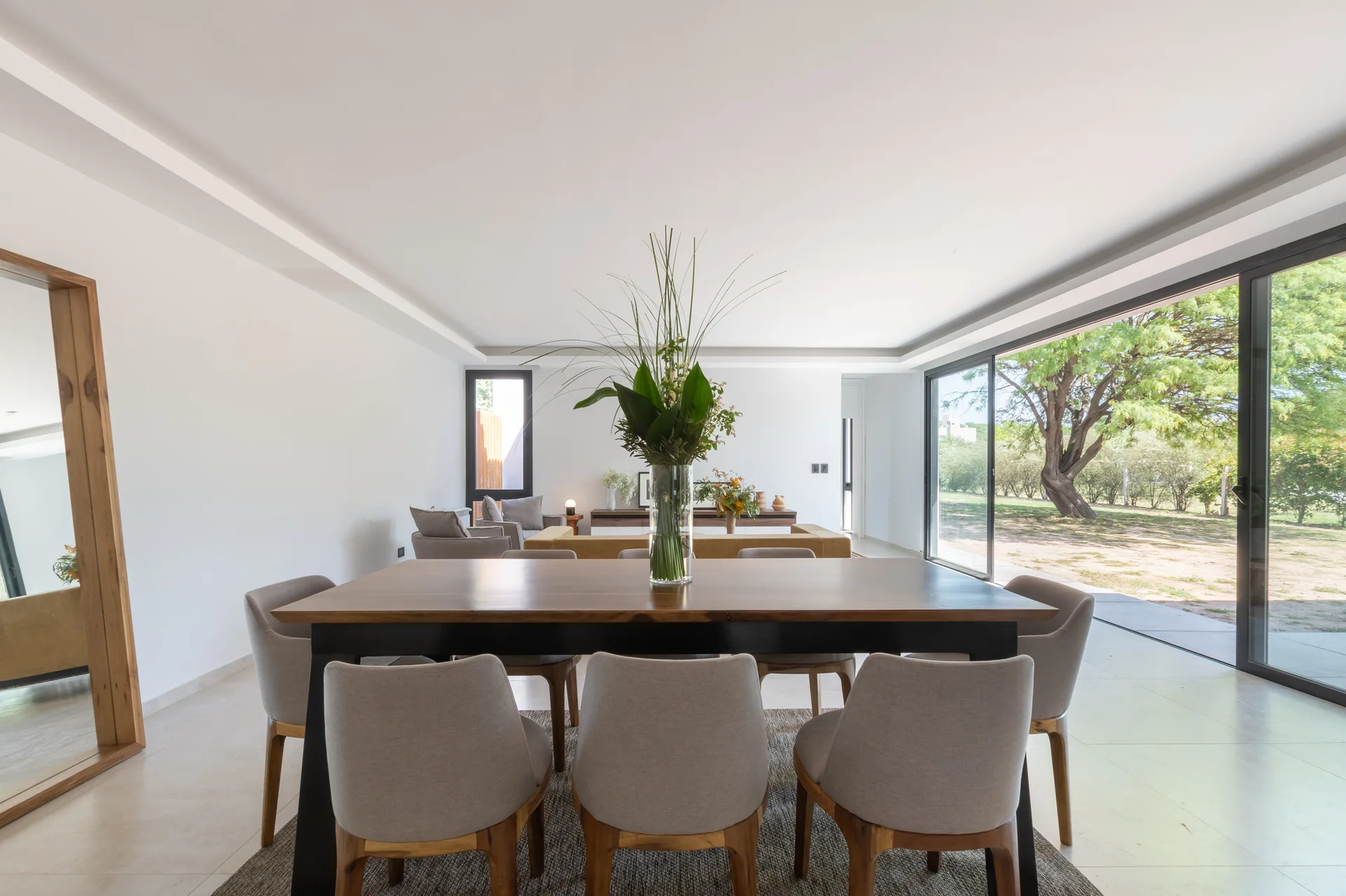 A large wooden dining table surrounded by grey upholstered chairs sits on a neutral rug before sliding glass doors opening to a green lawn.