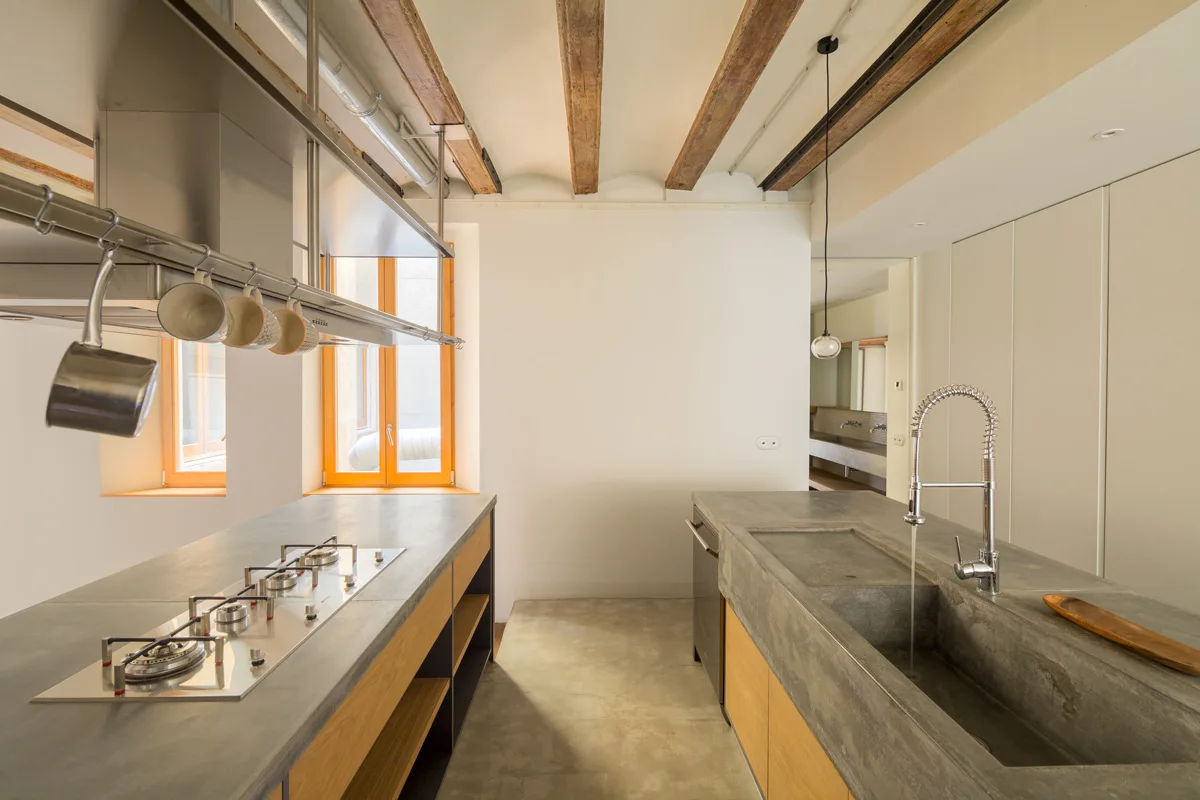 A modern kitchen features two concrete-topped islands, a stainless steel faucet, and exposed wooden ceiling beams over a concrete floor.