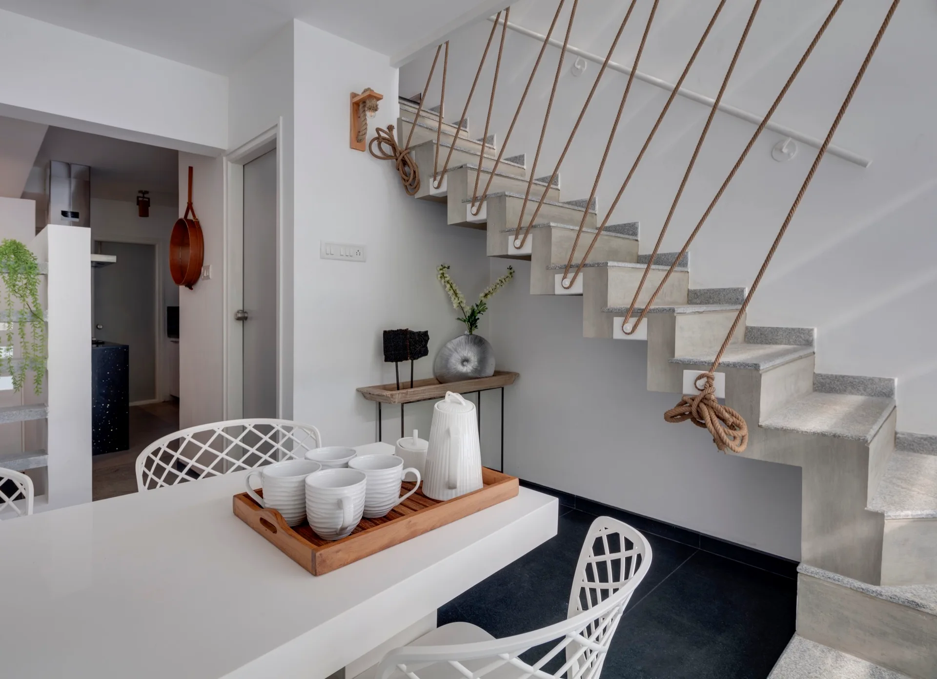 A modern dining room features a white table with lattice-back chairs next to a concrete staircase with a thick natural rope railing.