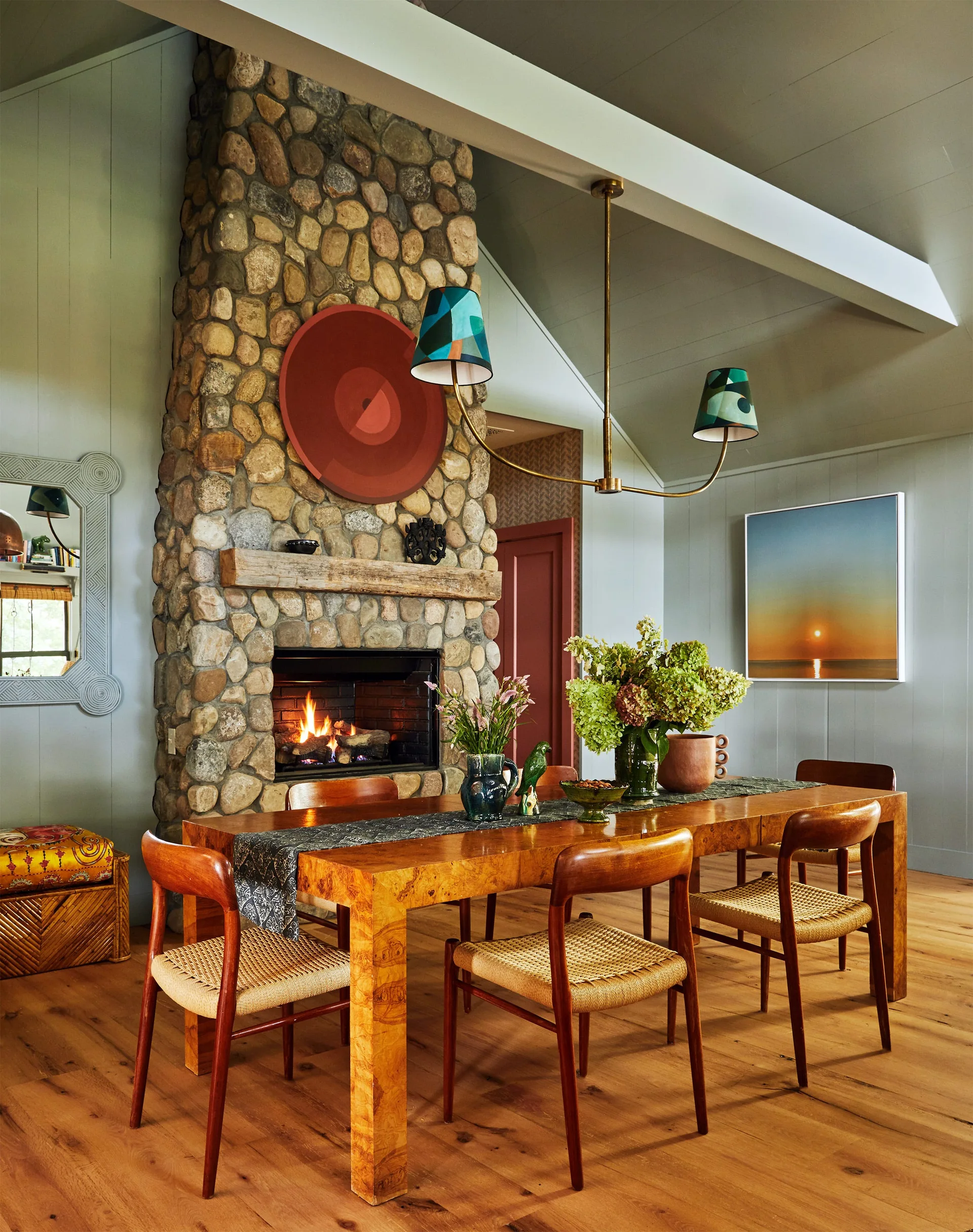 A dining room features a burl wood table, woven-seat wooden chairs, a large stone fireplace, and a brass chandelier with patterned green shades.