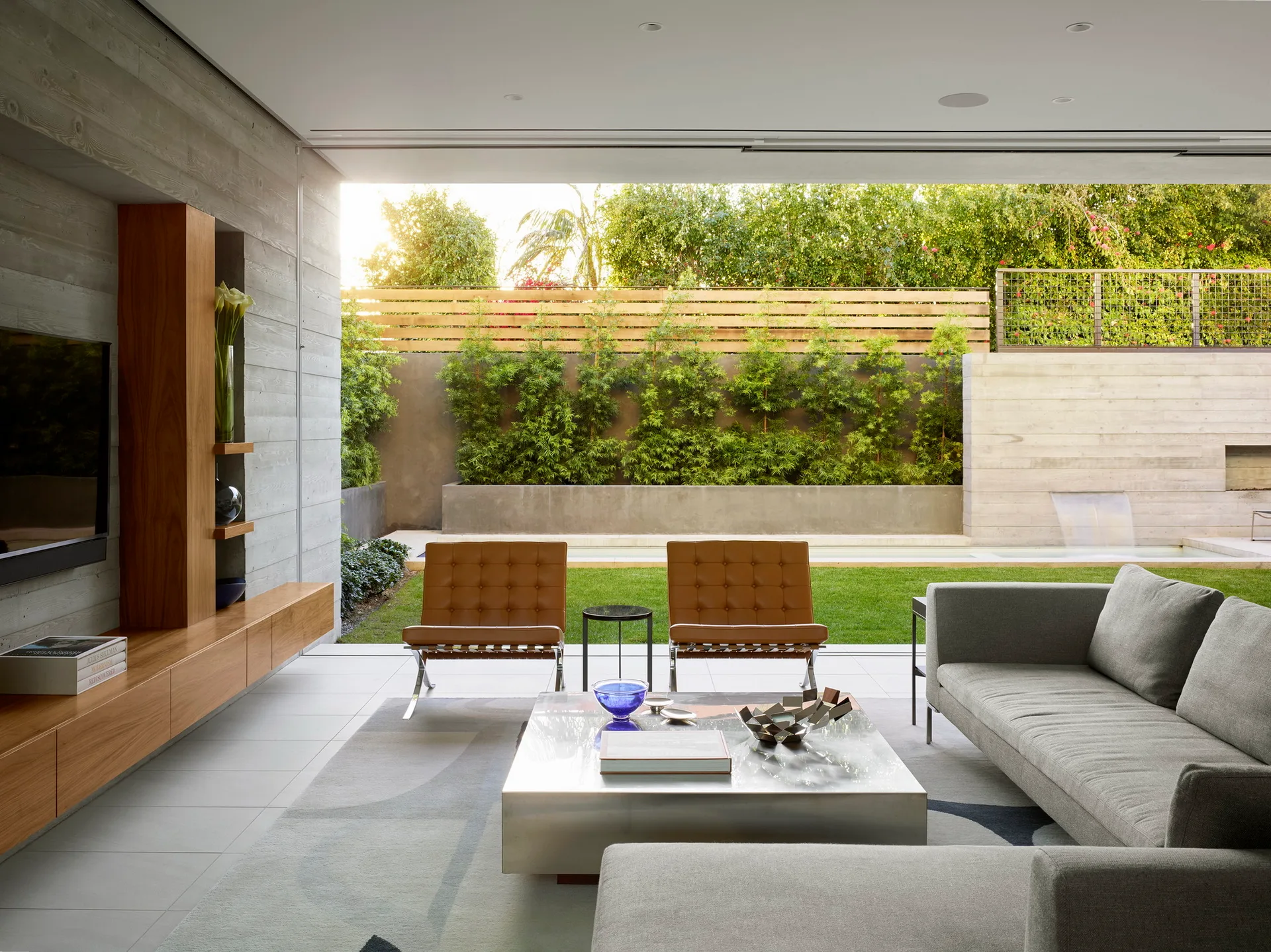 A minimalist living room with a grey sofa, tan leather chairs, and a floating wood console opens to a landscaped garden patio.
