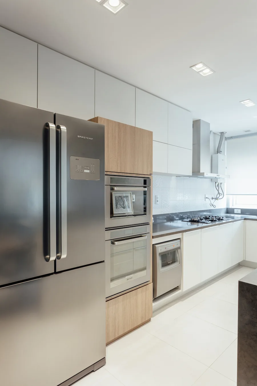 A modern kitchen with white flat-panel upper cabinets, a light wood column enclosing built-in ovens, and a stainless steel refrigerator.