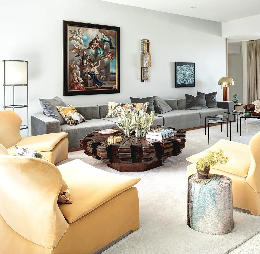 A contemporary living room with a long grey sofa, two yellow velvet armchairs, a carved round wooden coffee table, and a large framed classical painting.