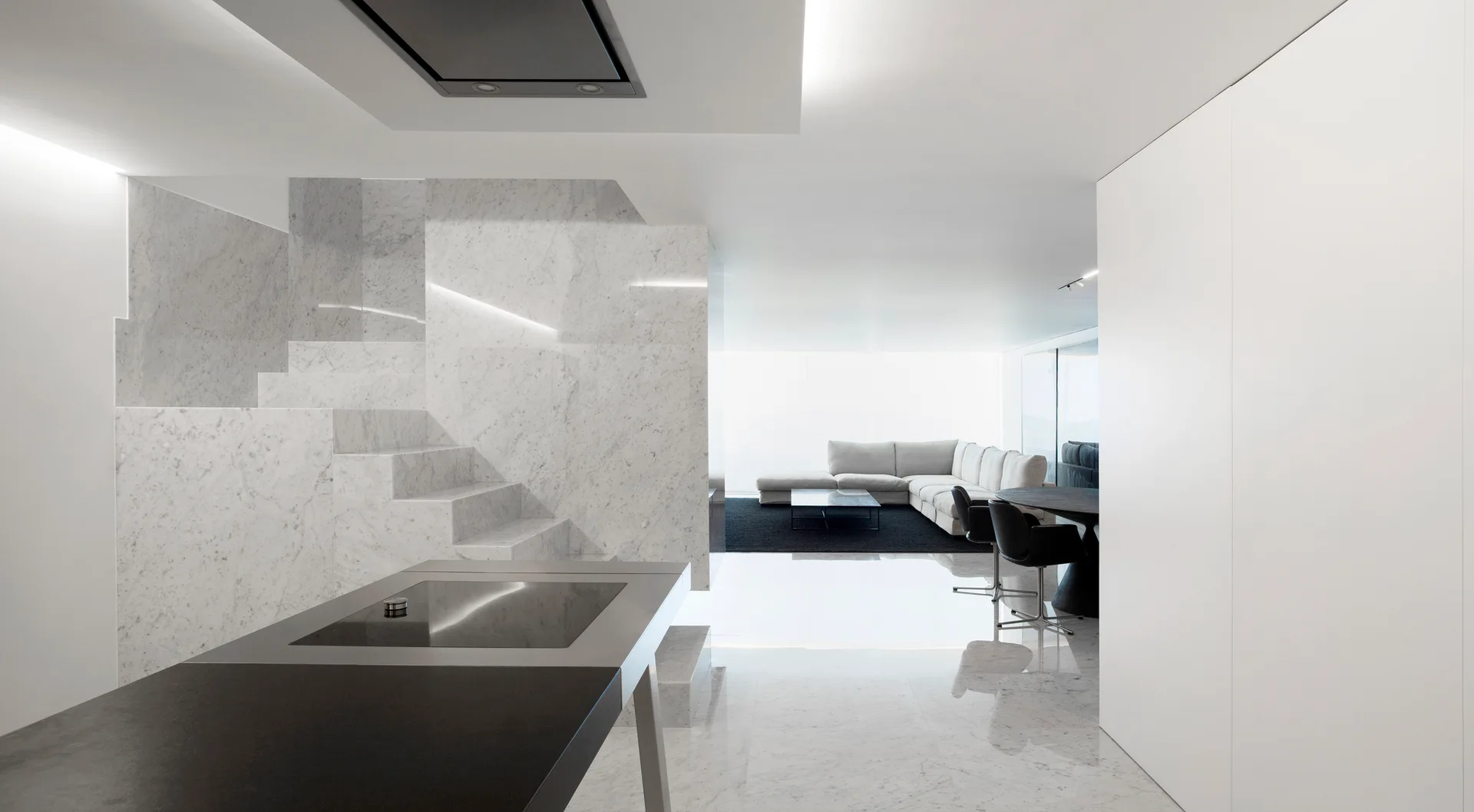 A minimalist open-plan living space features a polished marble floor, a solid marble staircase, a dark kitchen island, and a distant white sectional sofa.