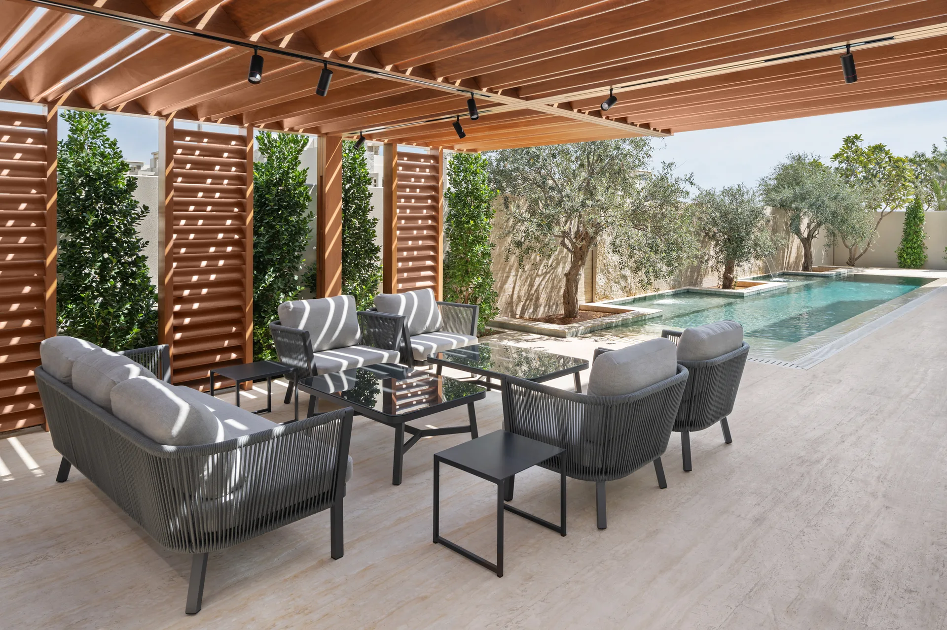 Gray outdoor seating with woven rope backs sits under a wood slatted pergola next to an in-ground swimming pool on a beige stone patio.