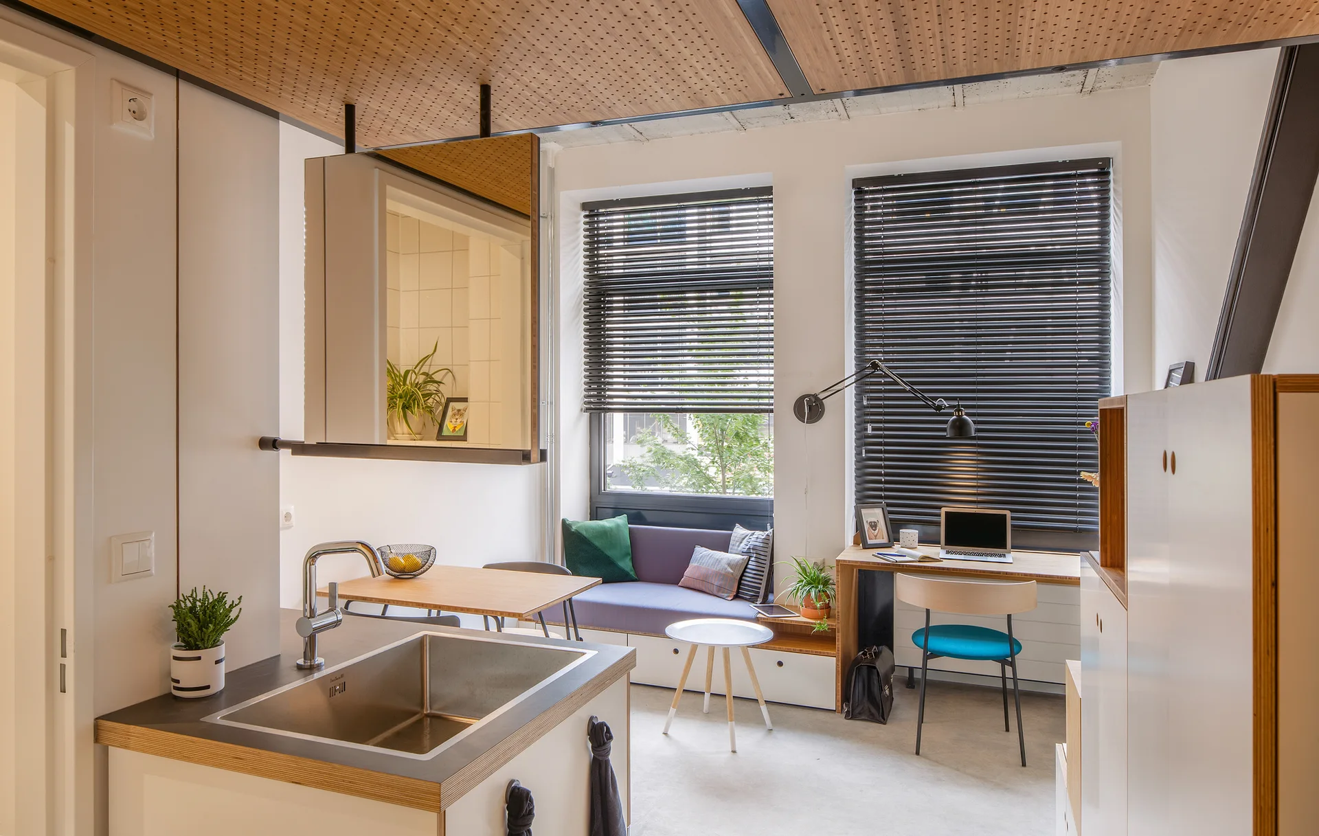 A studio apartment interior featuring a kitchen sink island, a daybed under a window with black blinds, a built-in wooden desk, and a perforated ceiling.