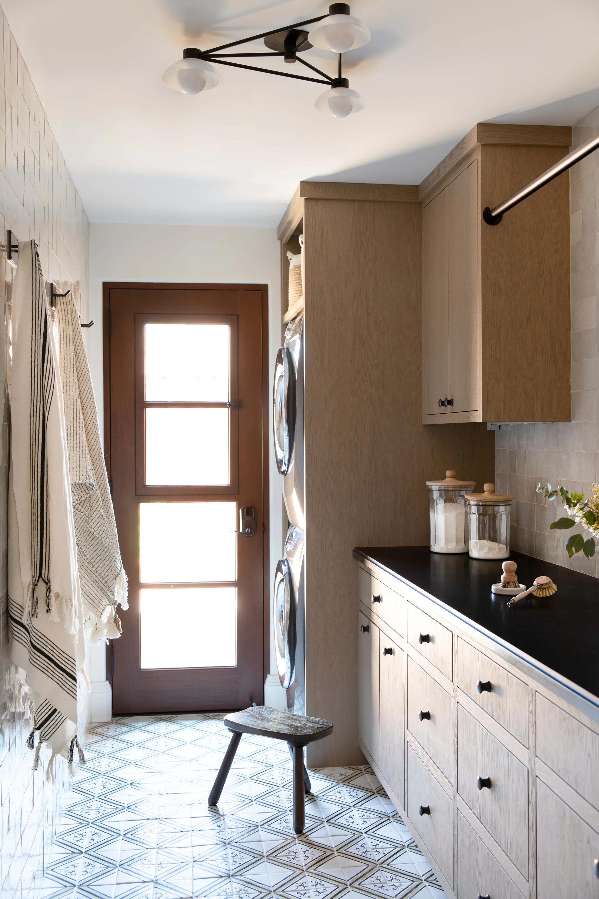 A laundry room with light wood cabinets, stacked appliances, black countertops, and geometric floor tiles leading to a glass paneled wood door.