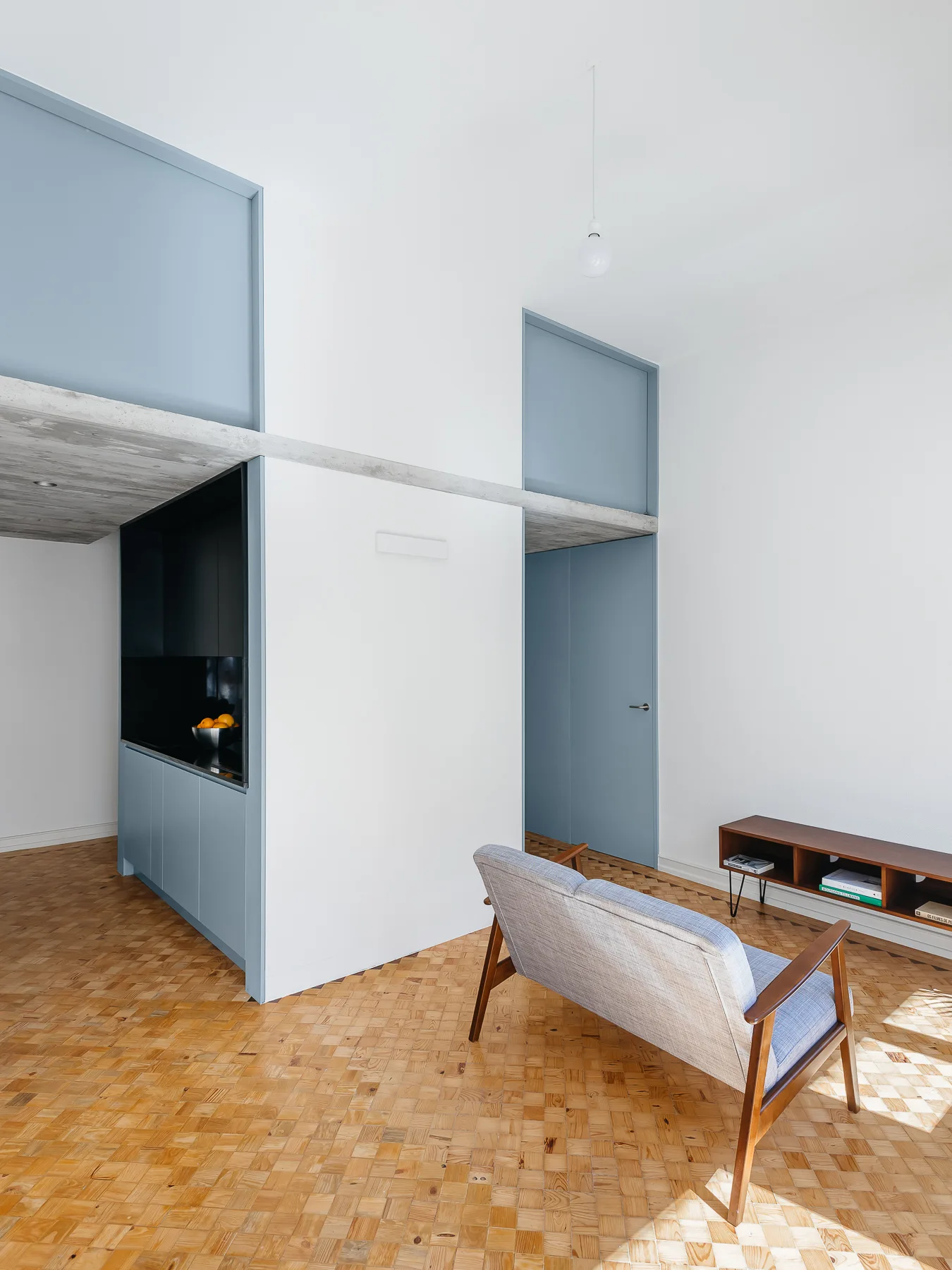 A minimalist living space featuring a wood block parquet floor, a concrete support beam, a mid-century loveseat, and powder blue doors and cabinets.