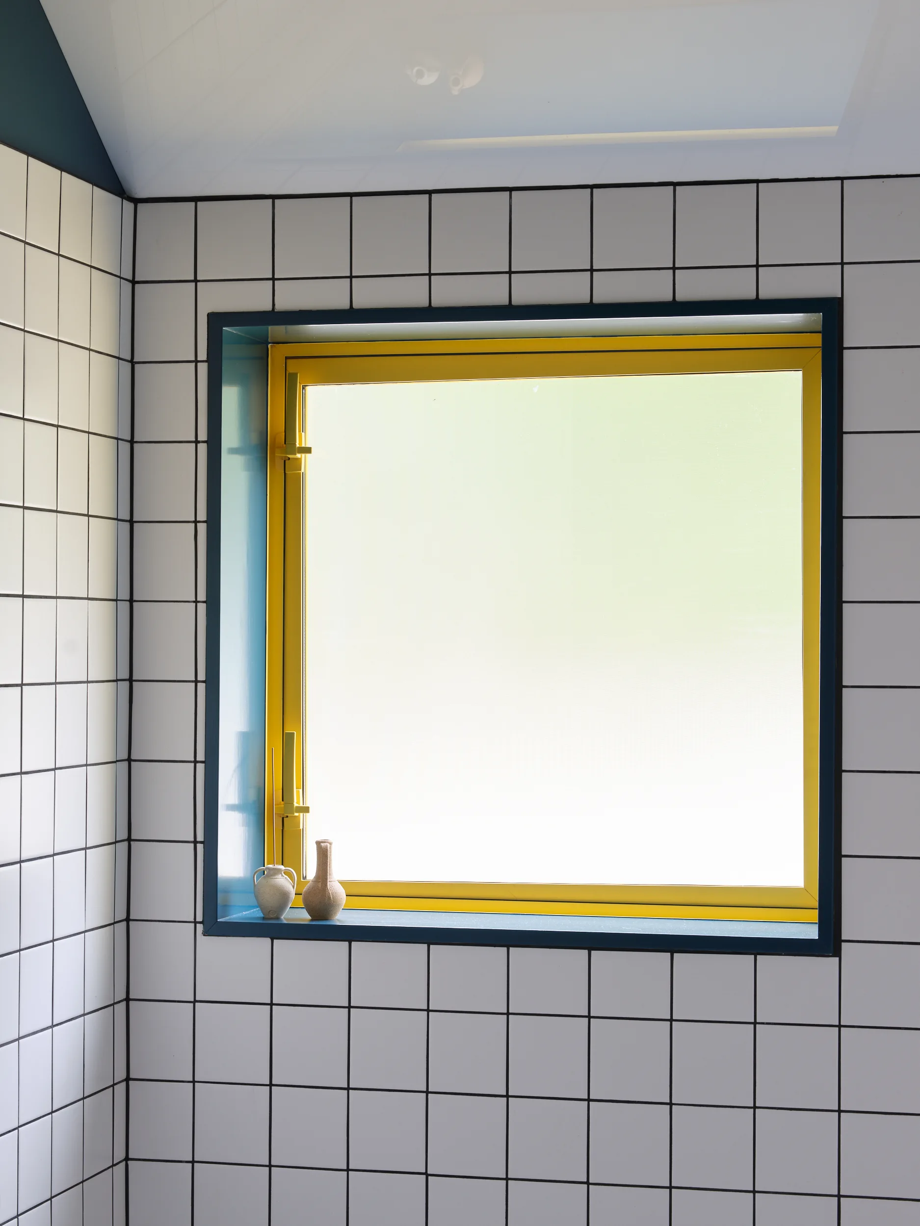 A recessed square window features a yellow frame and dark blue trim set in a white tile wall with black grout.