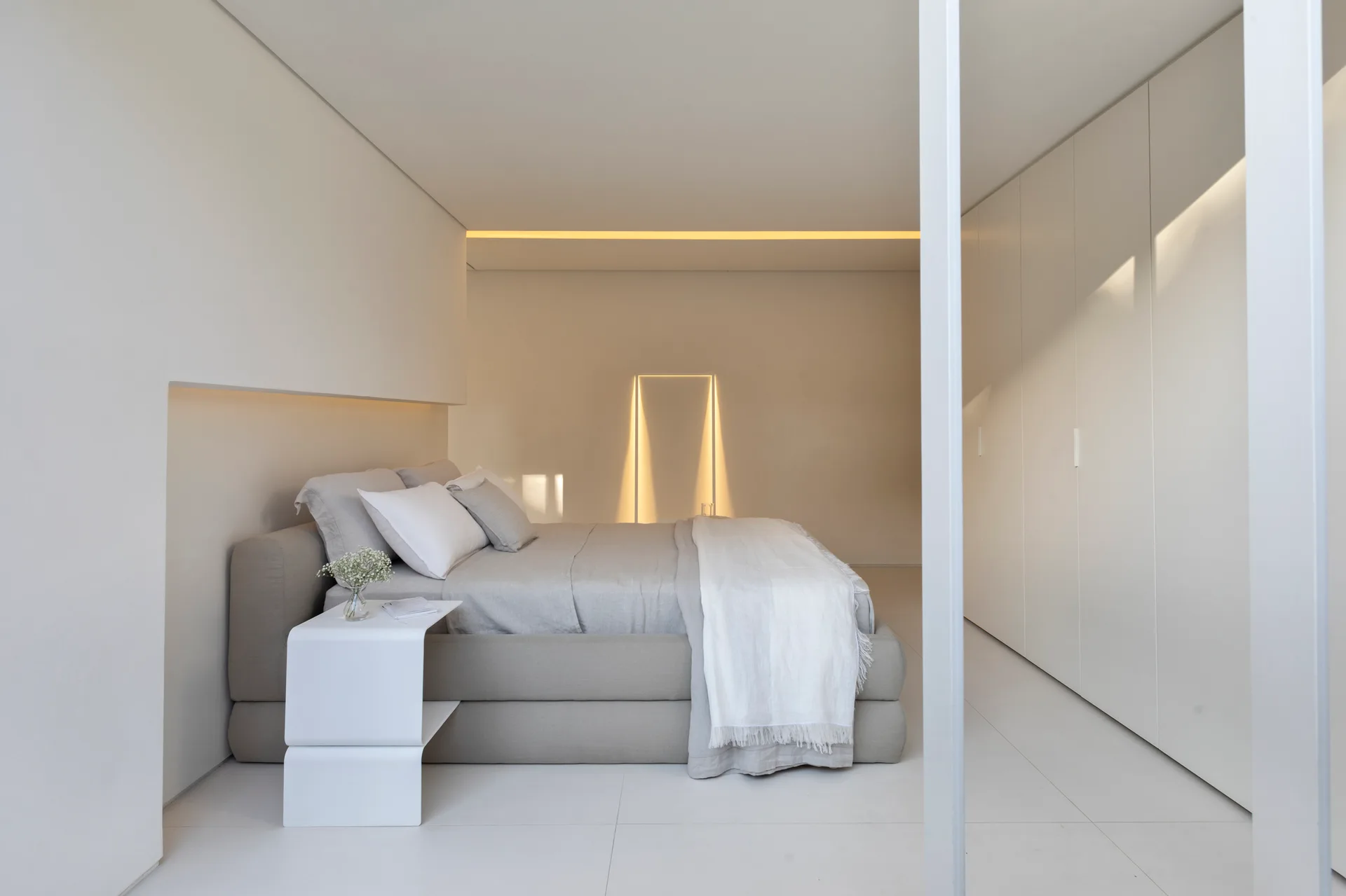 A minimalist bedroom features an upholstered platform bed, a seamless floor-to-ceiling white wardrobe, and integrated warm cove lighting.