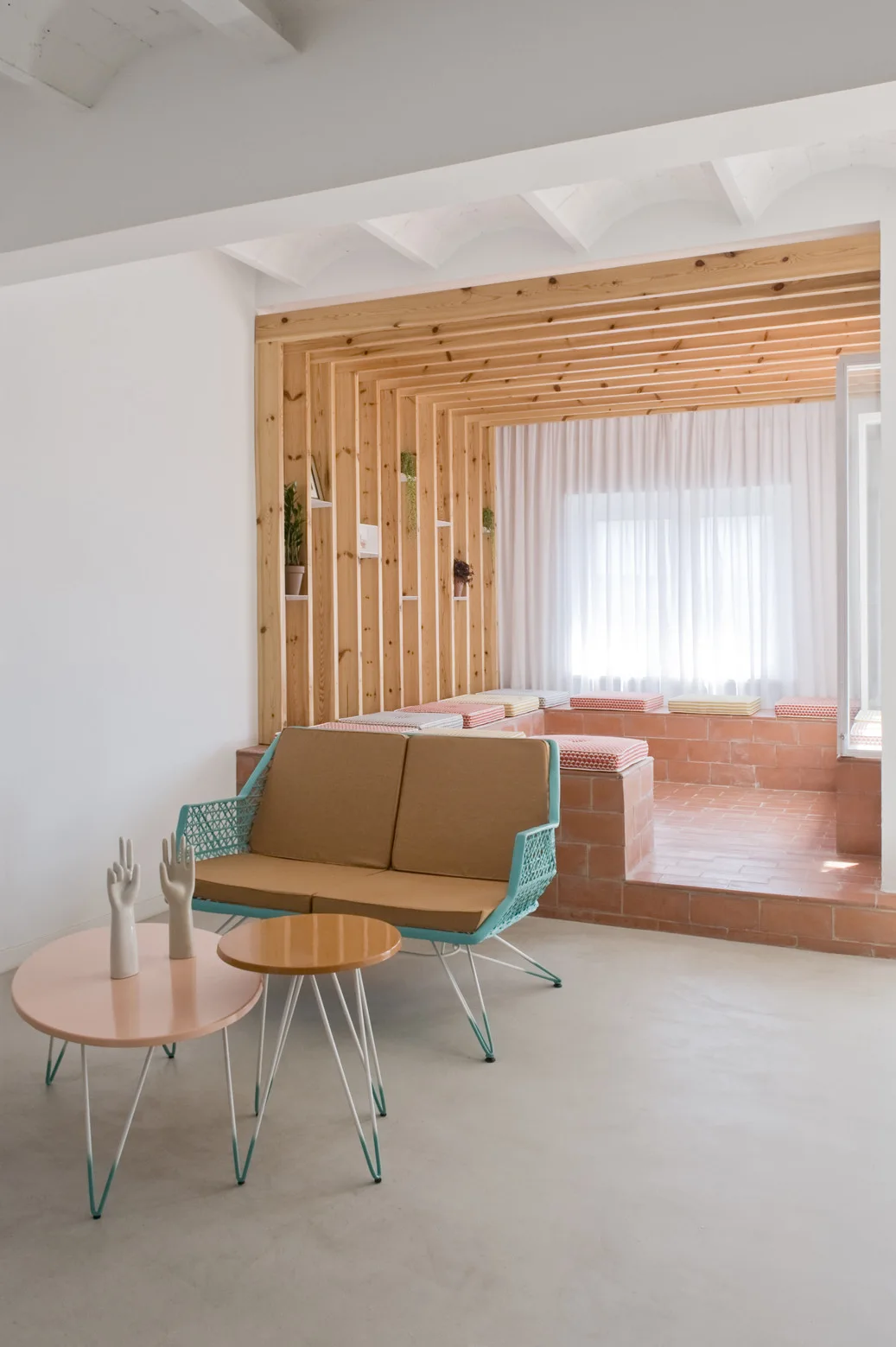 A modern lounge features a turquoise frame sofa, pink coffee table, and a raised terracotta brick platform framed by vertical wood slats.