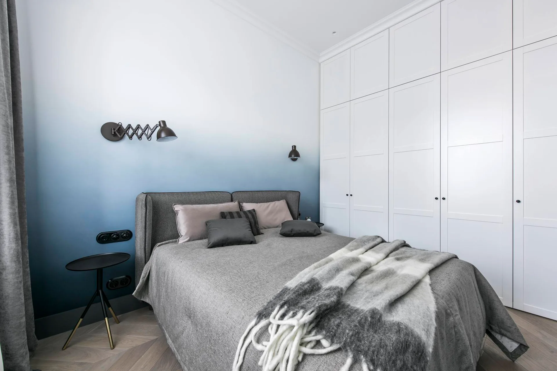 A contemporary bedroom features a bed with grey bedding set against a blue-and-white gradient accent wall next to built-in white wardrobes.