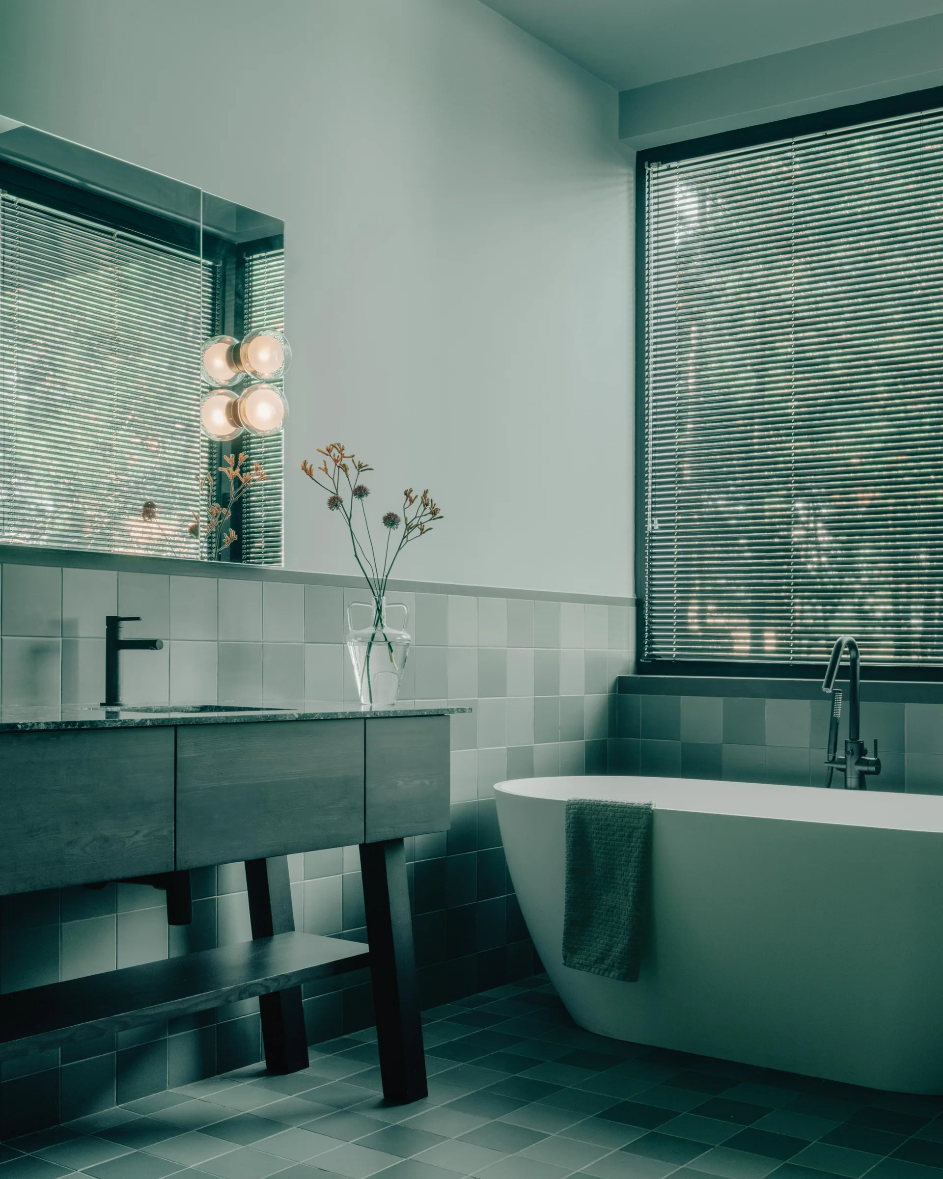 A modern sage green bathroom features a dark wood vanity, a white freestanding bathtub under a window with blinds, and square grid tiling on the wall.