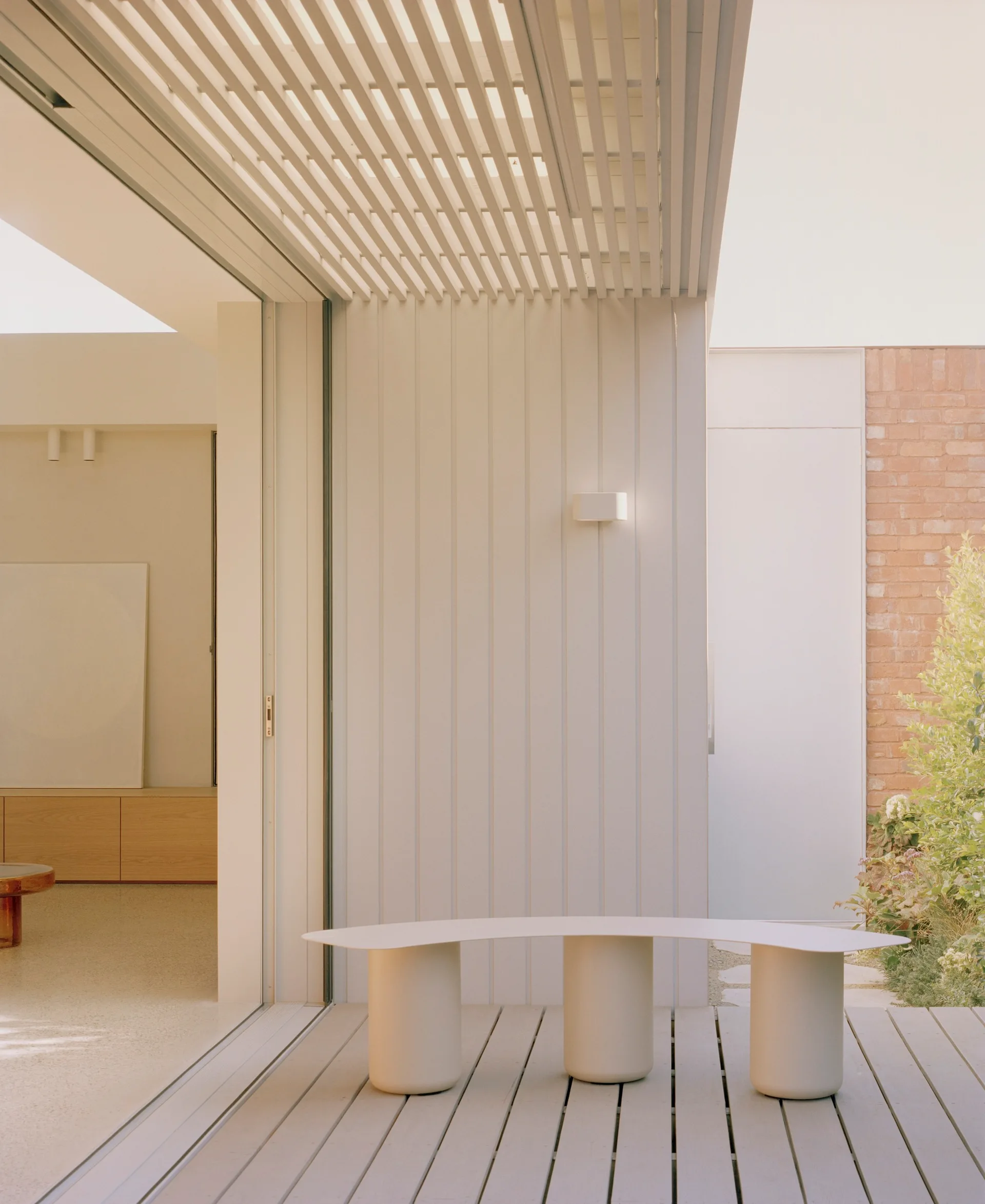 A curved cream-colored outdoor bench stands on a grey wooden deck underneath a slatted white pergola ceiling.