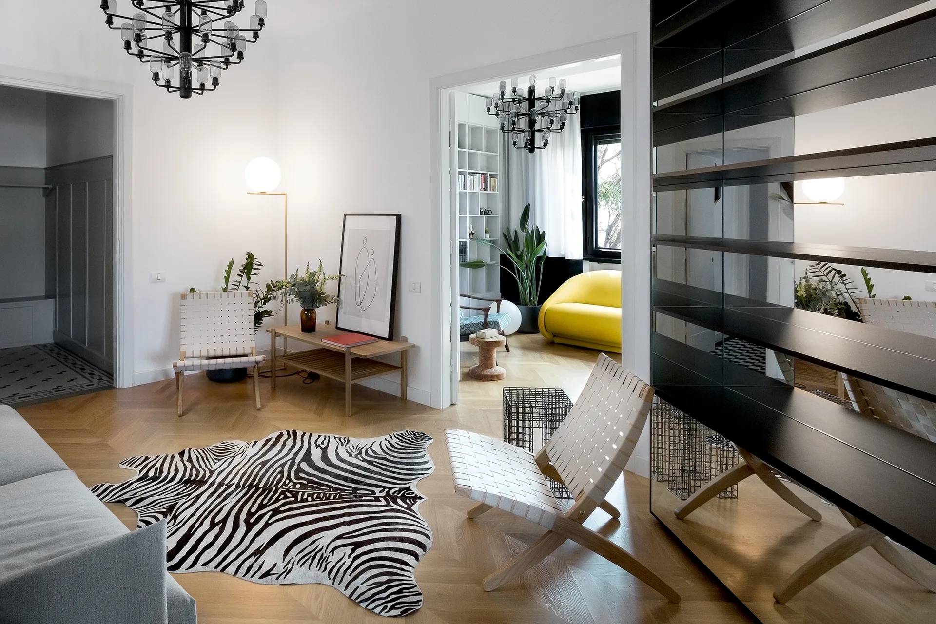 A bright living room features light herringbone wood floors, woven strap lounge chairs, a zebra-patterned rug, and a large black shelving unit.