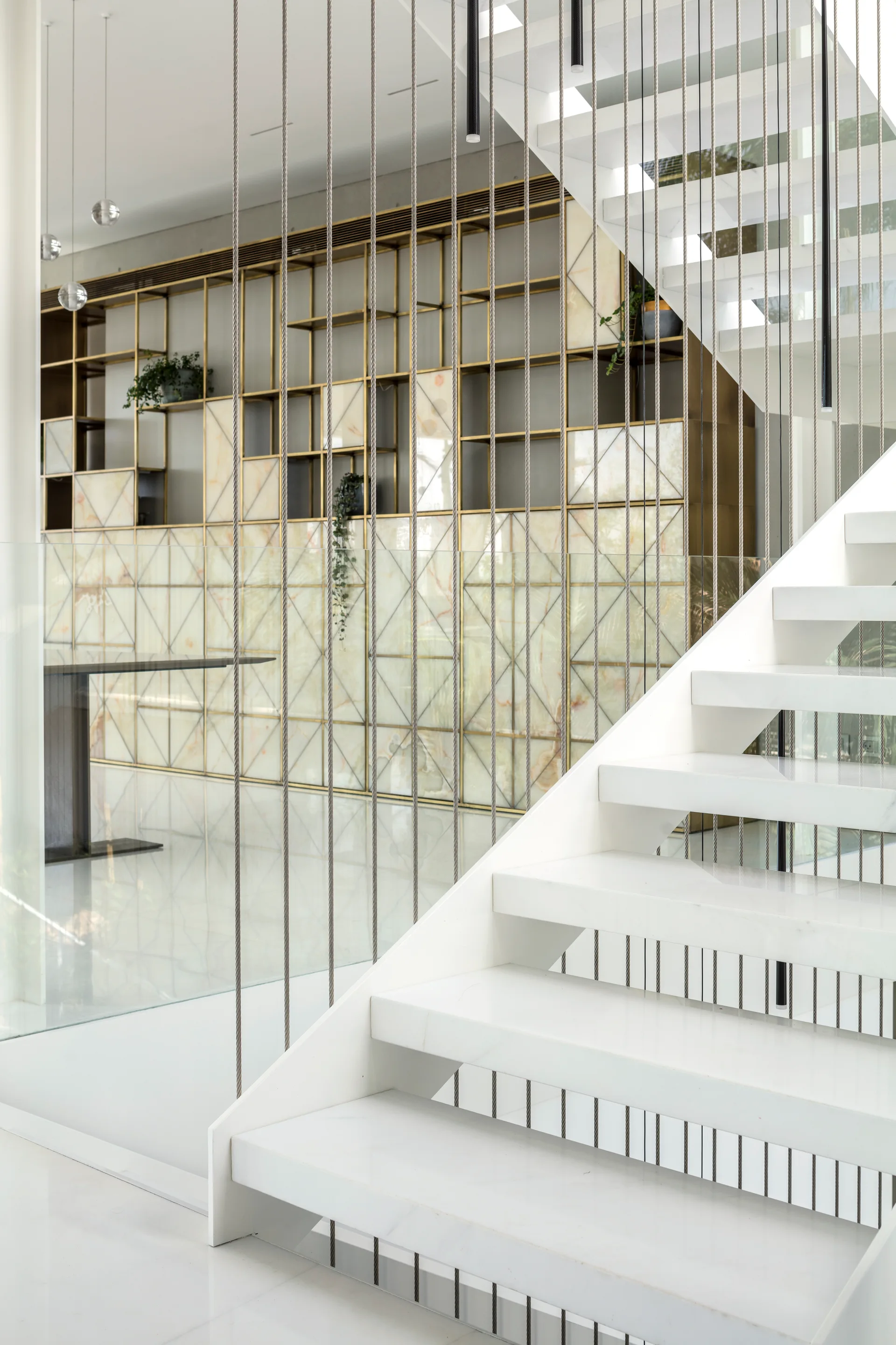 A white floating staircase features vertical steel cable railings next to a gold metal shelving unit with marble-patterned backing panels.