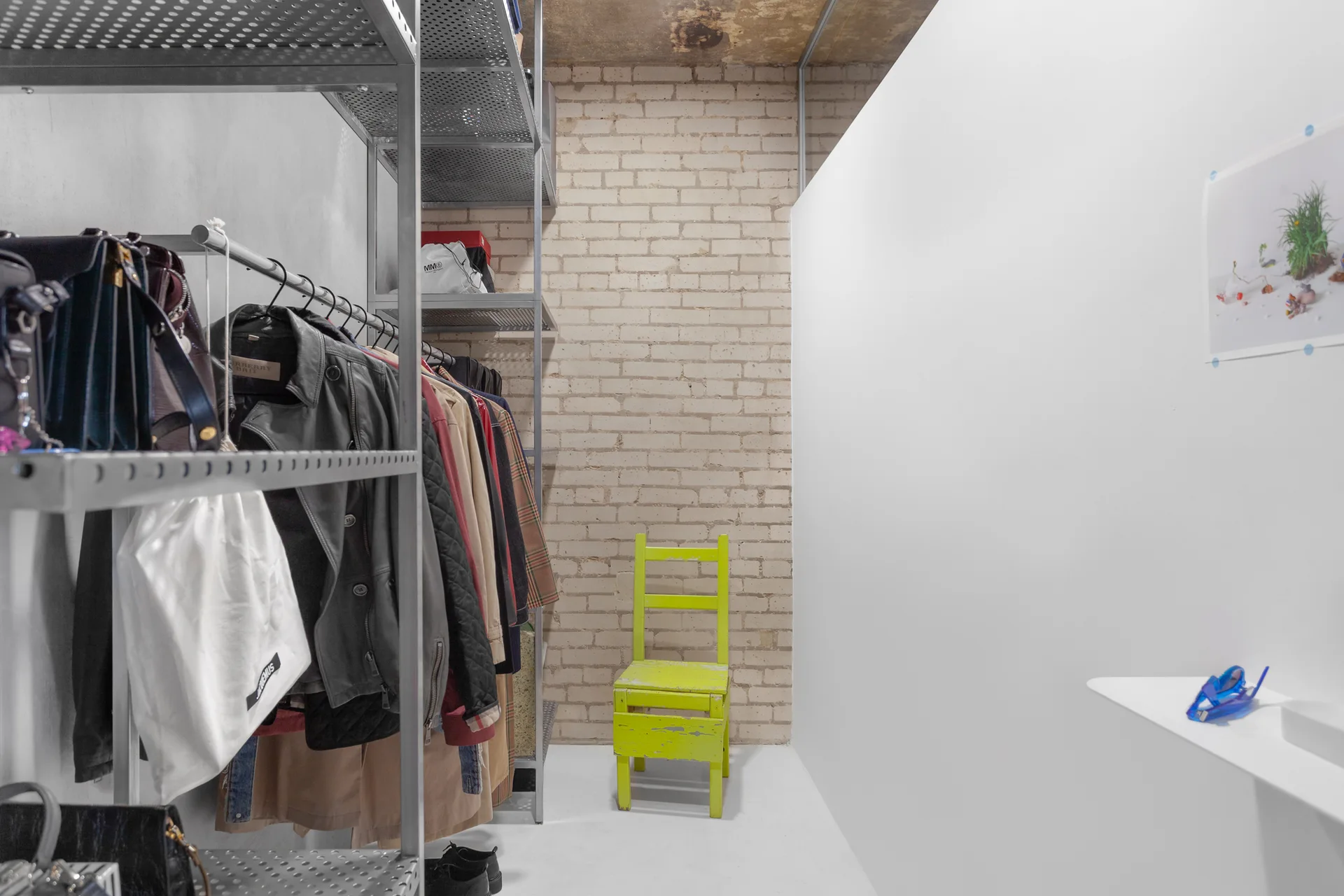 An industrial closet features perforated metal clothing racks and shelves alongside an exposed brick wall and a bright yellow wooden chair.